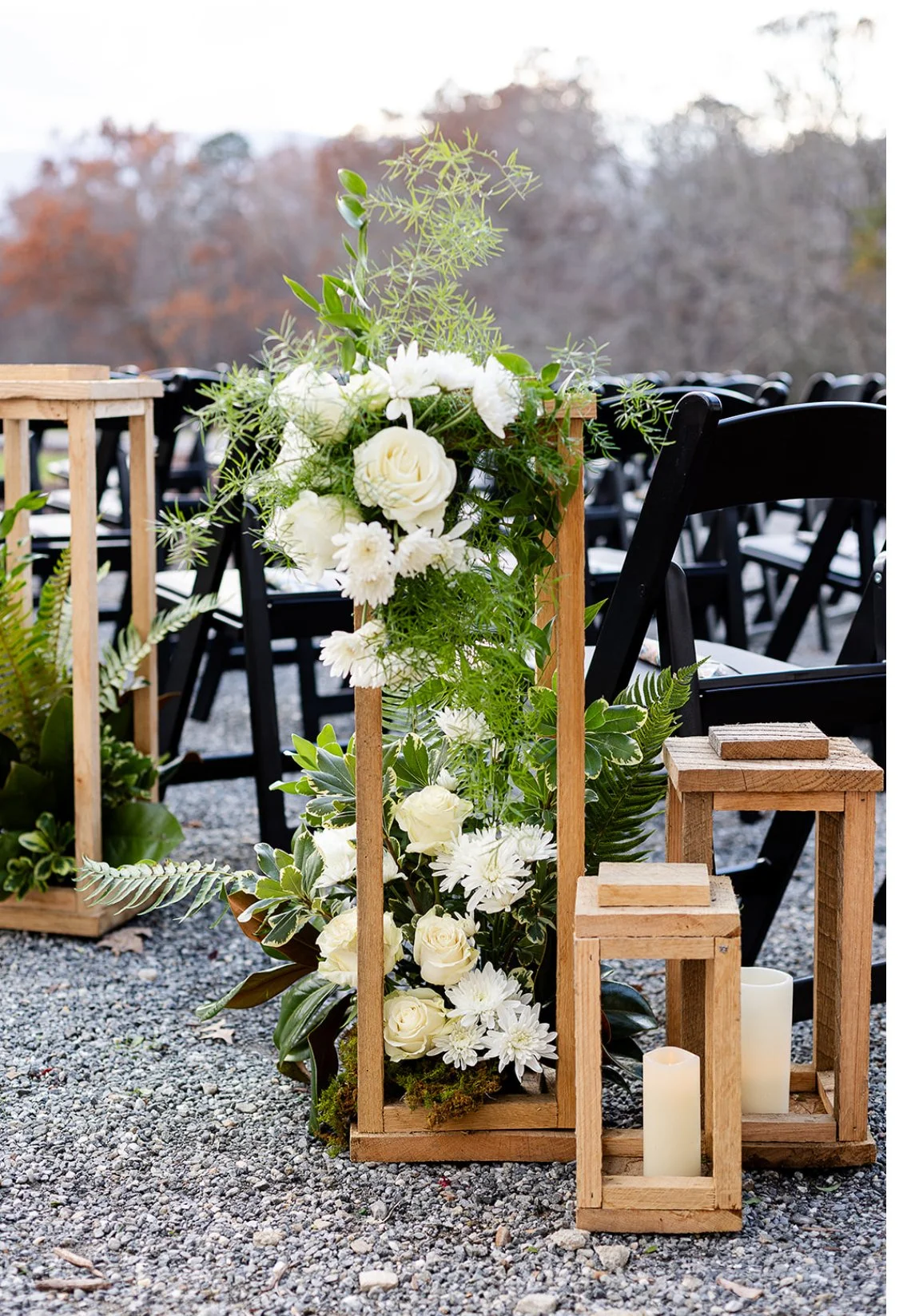 Floral arrangements with white flowers and green foliage in wooden frames, with candles on gravel ground, set outdoors with black chairs in the background.