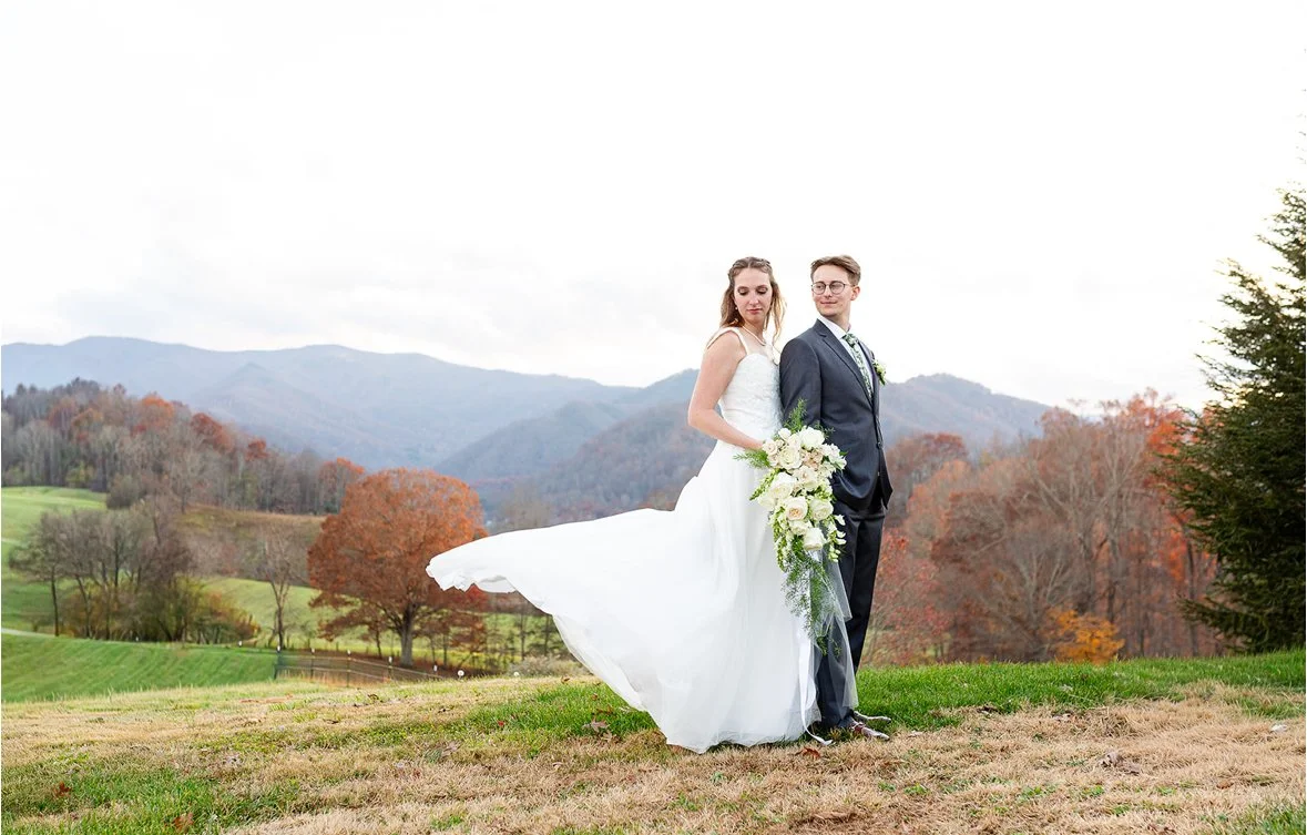 A bride and groom standing outdoors on a grassy field with mountains and autumn trees in the background. The bride is in a white wedding dress holding a large bouquet, and the groom is in a dark suit with a tie.