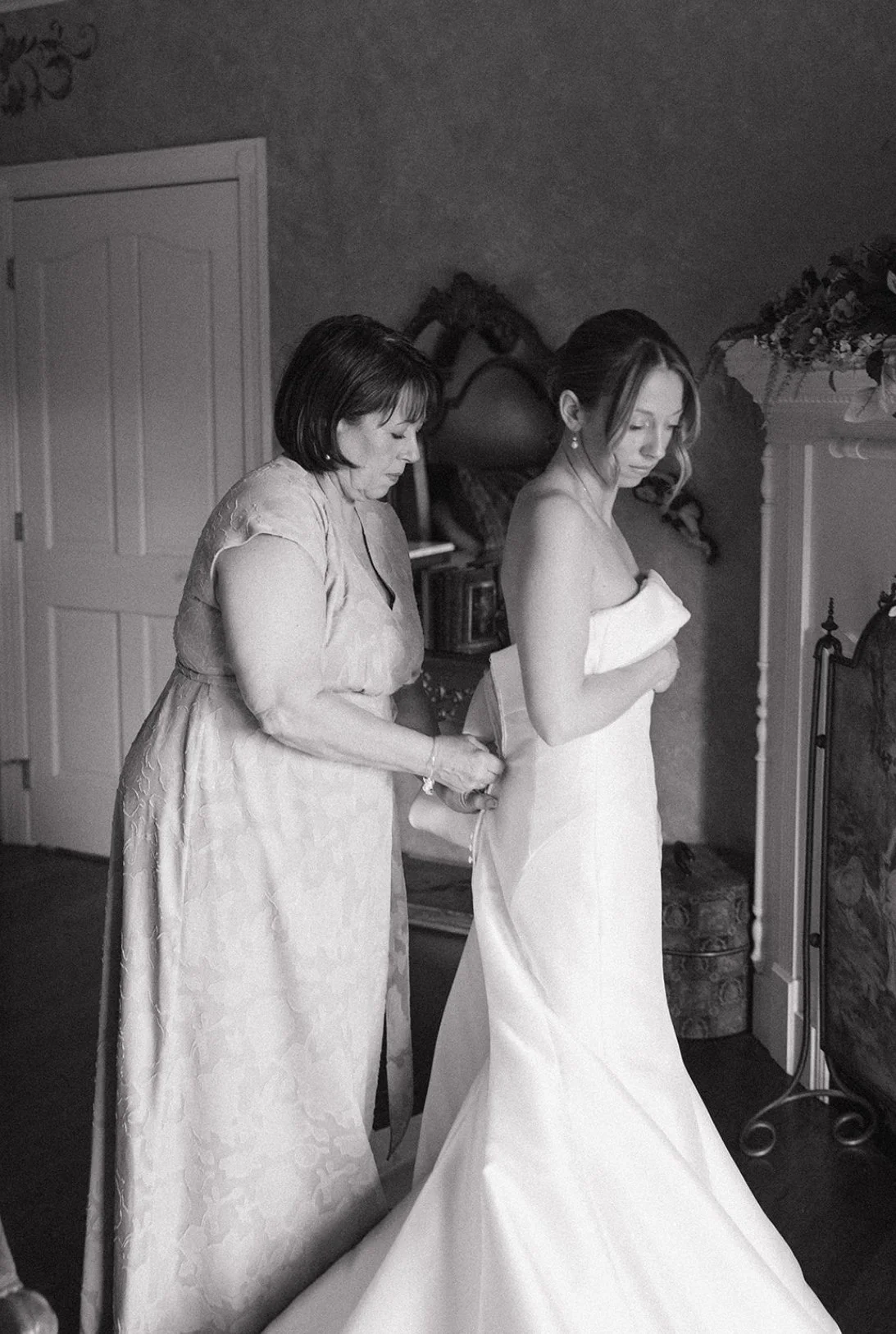 A woman in a wedding dress with a strapless gown, standing in a room as another woman, likely helping her with her dress, adjusts or fastens the back of her gown. The room has a fireplace and decorative elements.