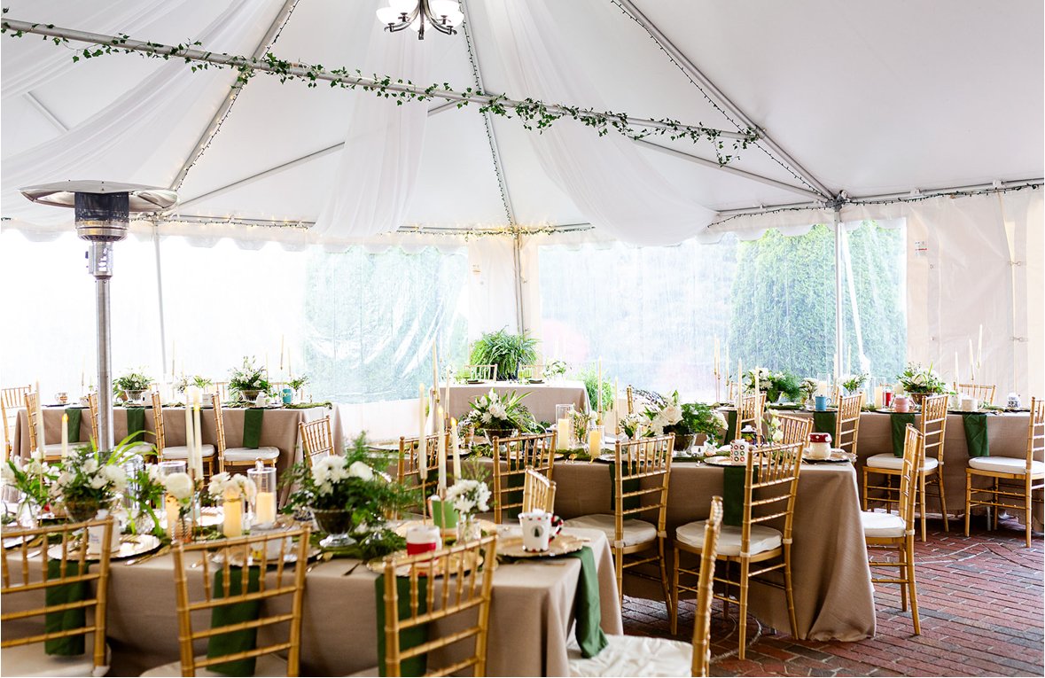 Indoor event space decorated for a celebration with tables, gold chairs, white flowers, candles, and greenery under a white tent