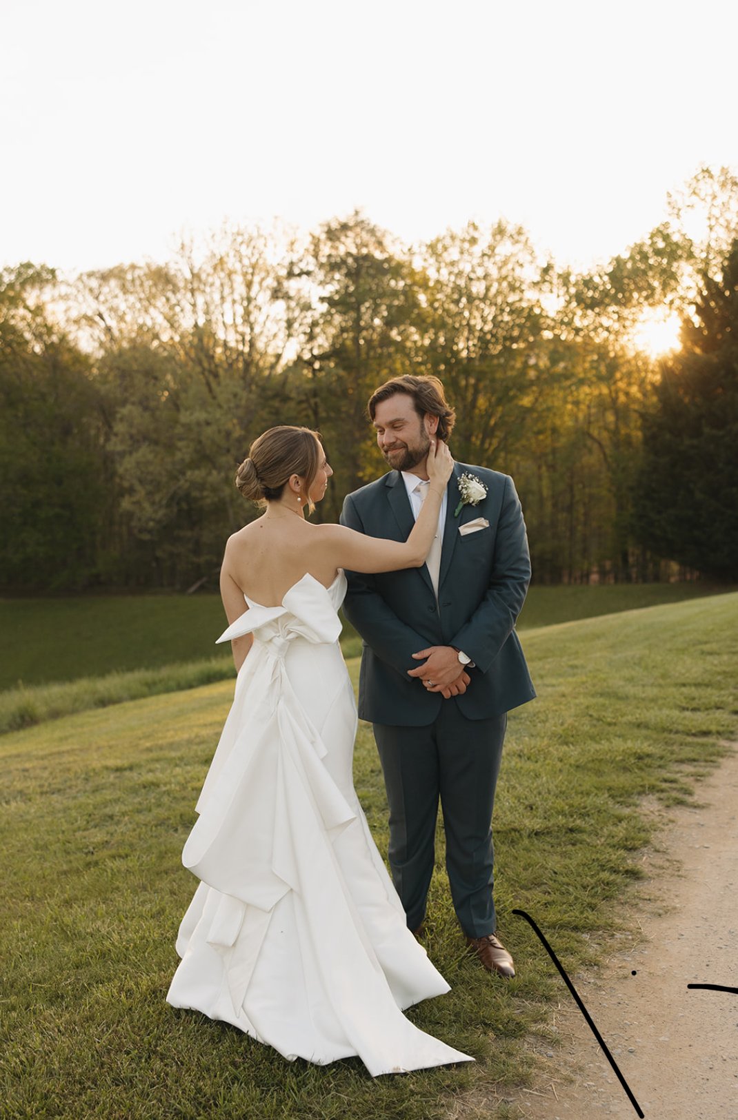 A bride and groom standing on grass outdoors at sunset, with the bride touching the groom's face and both smiling.
