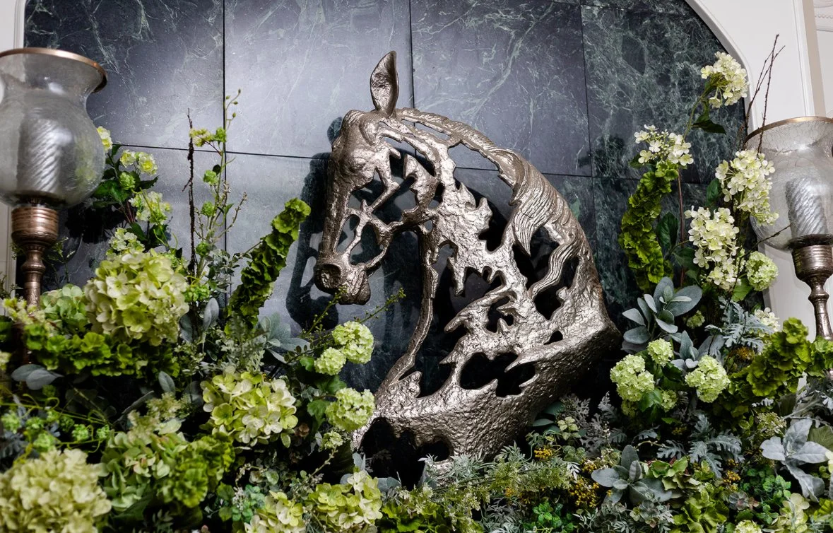 A decorative metal horse head sculpture surrounded by green and white floral arrangements.