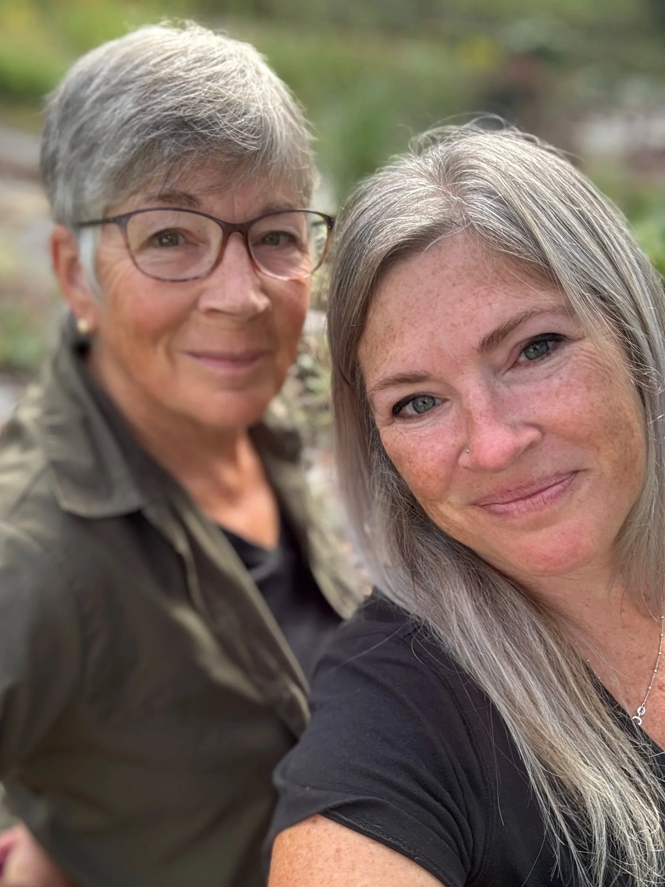 Two women taking a selfie outdoors with greenery in the background. The woman on the left has short gray hair, glasses, and a green jacket. The woman on the right has long gray hair, freckles, and is wearing a black top.
