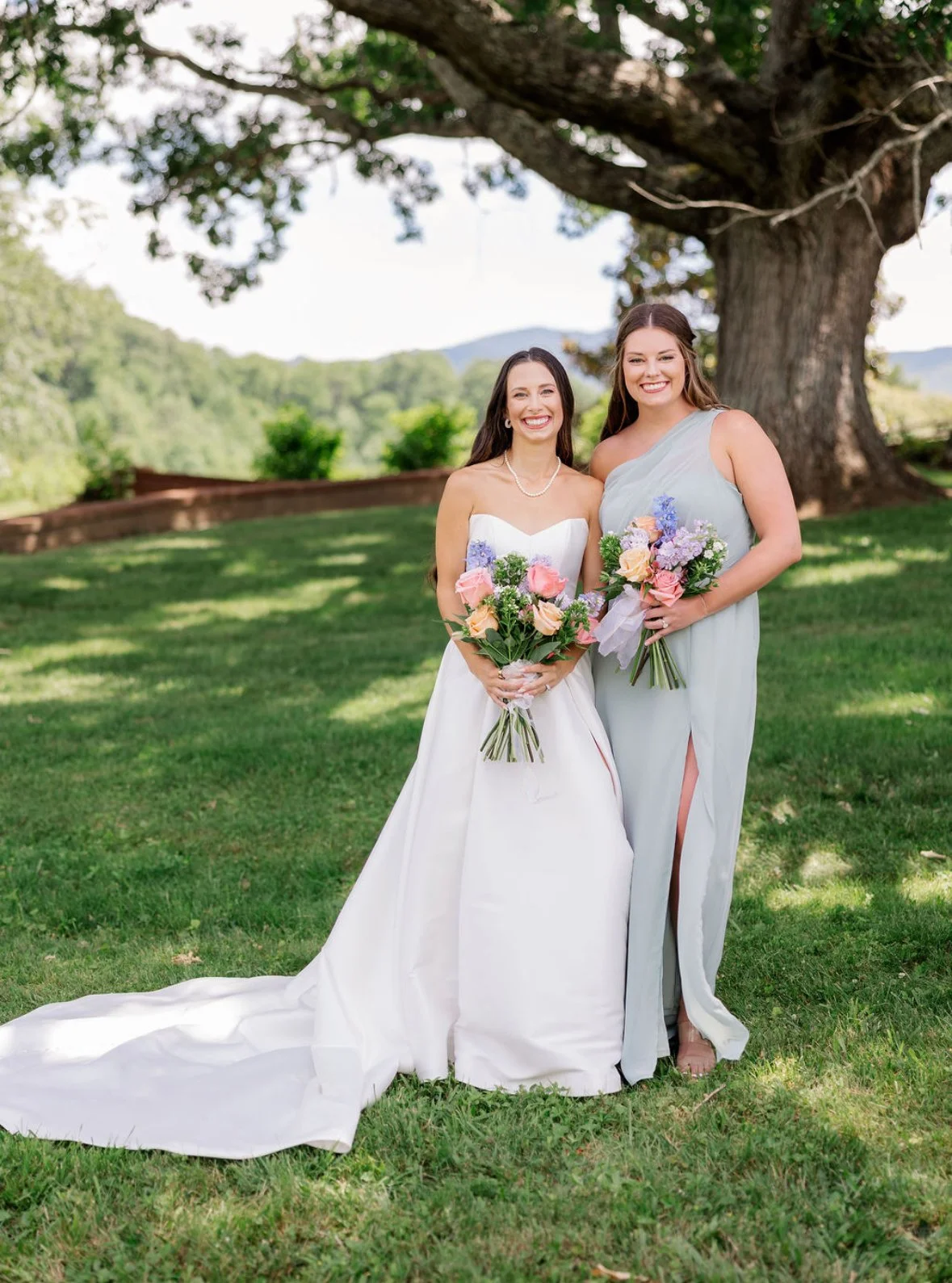Two women, one in a white wedding dress and the other in a light blue dress, holding bouquets, standing outdoors under a large tree with lush green grass and hills in the background.