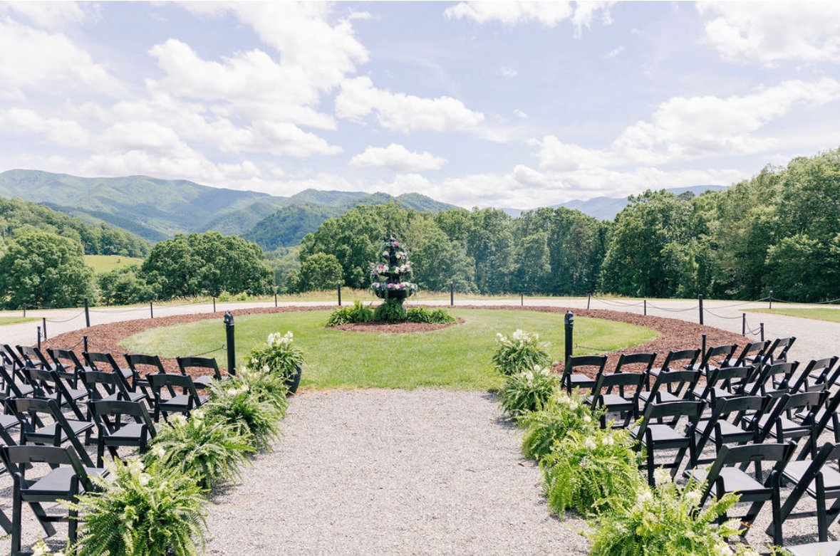 Outdoor wedding ceremony setup with black chairs arranged on both sides of a gravel aisle leading to a central fountain with mountains in the background.