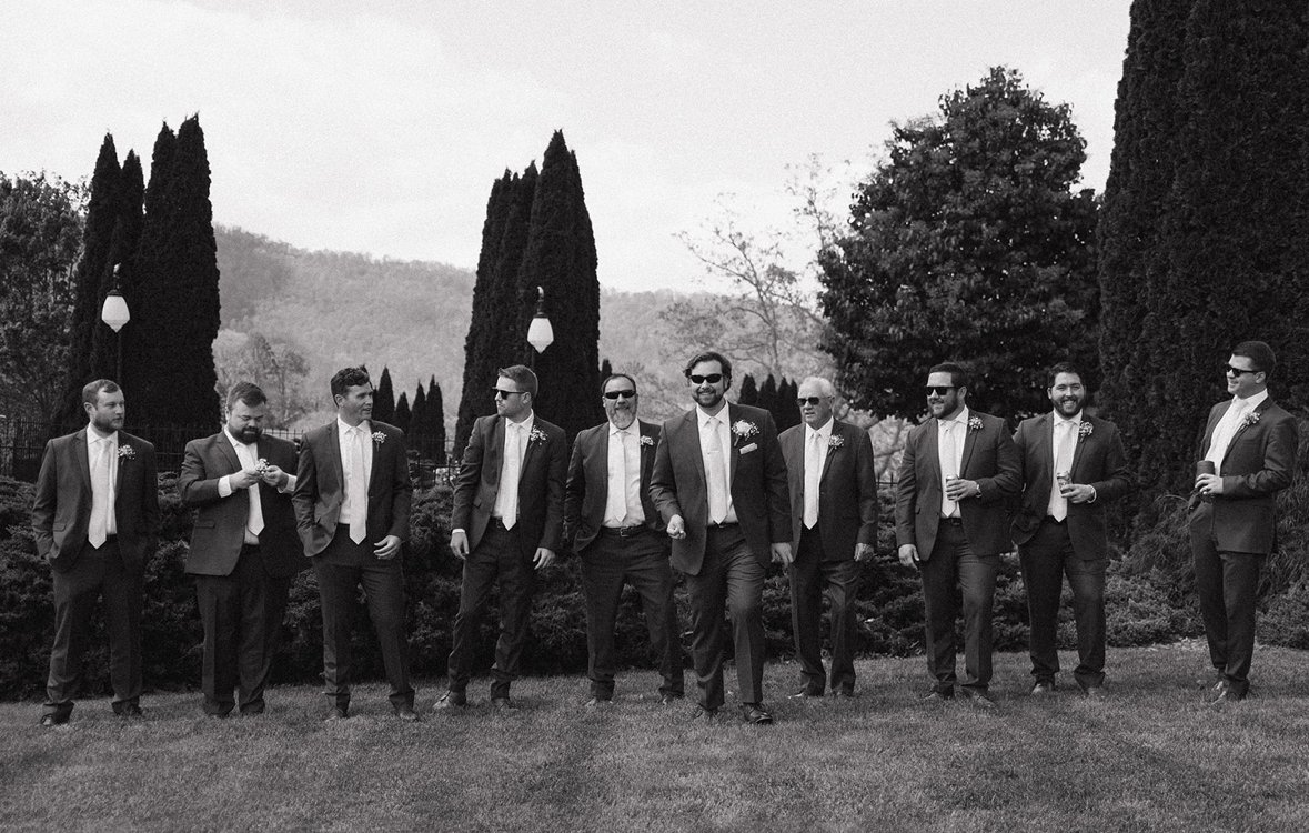 A group of men in suits walking outdoors in a park with tall trees and mountains in the background