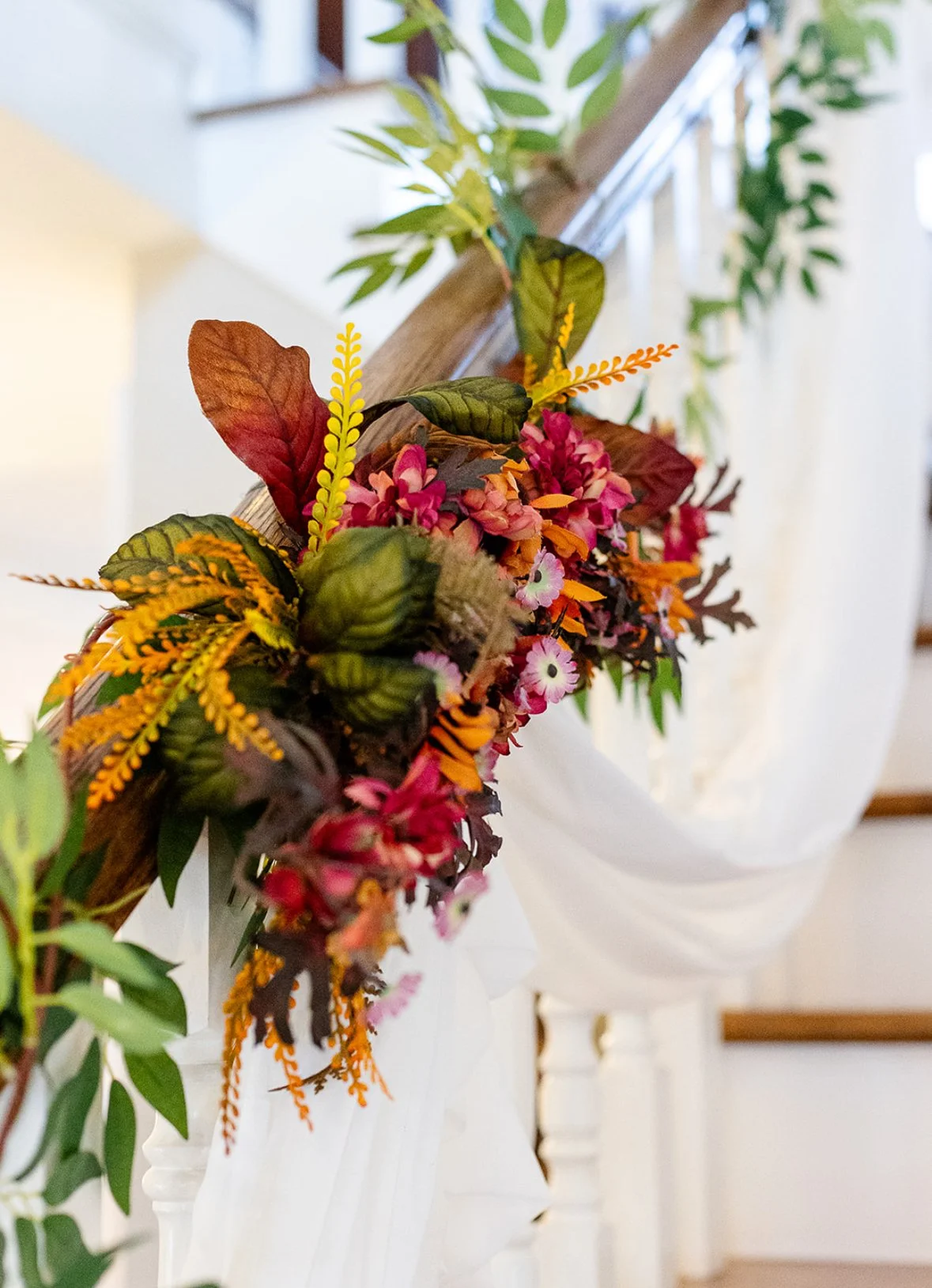 Floral garland with pink, orange, and green artificial flowers and leaves, draped along a wooden railing inside a house.