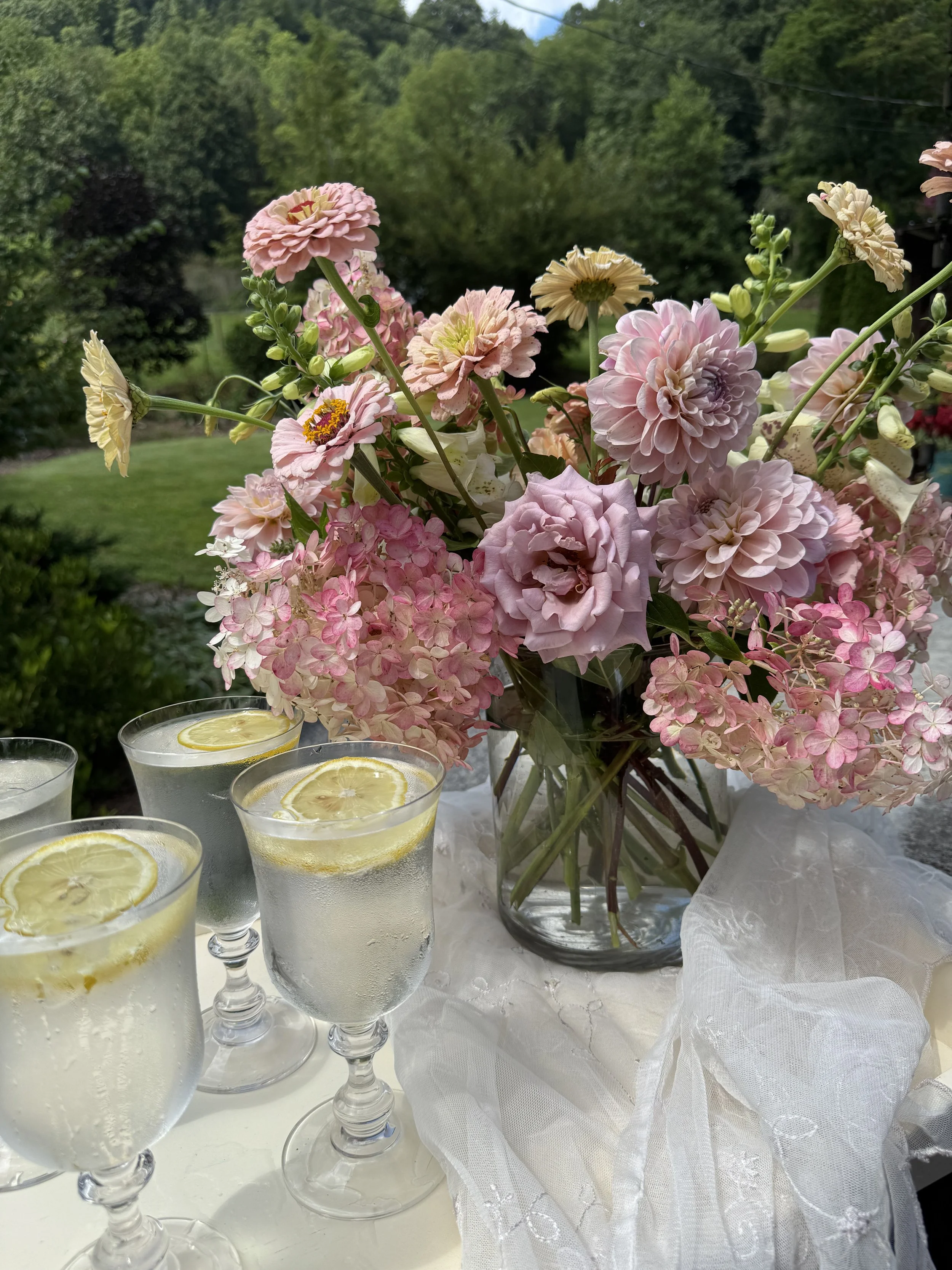 A bouquet of pink and cream flowers in a glass vase on a white lace tablecloth, with four glasses of lemonade with lemon slices, outdoors with green trees and a blue sky in the background.