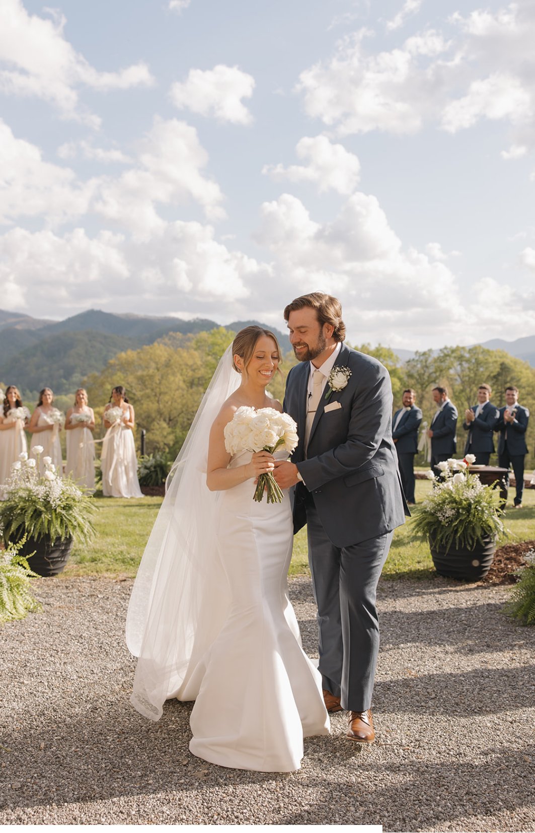 A bride and groom smiling as they hold hands during their outdoor wedding ceremony with mountain scenery in the background. Bridesmaids and groomsmen are visible in the background.