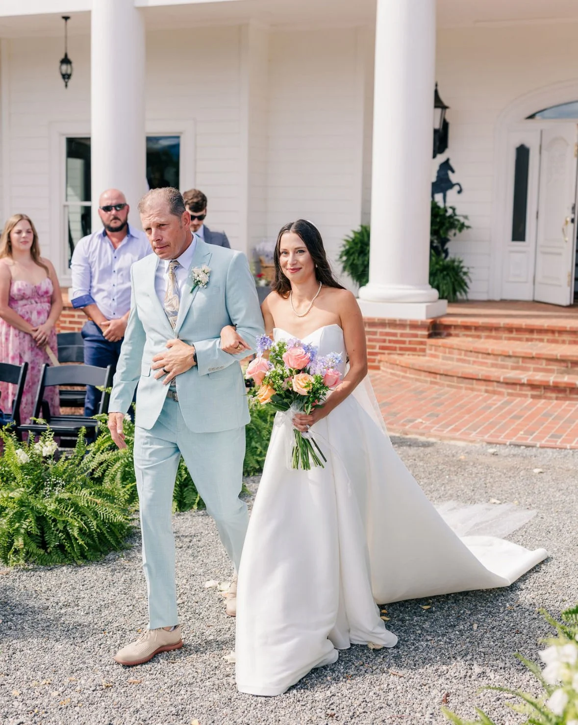 A bride in a white wedding dress being escorted down the aisle by a man in a light blue suit at an outdoor wedding ceremony, with guests and a white house in the background.