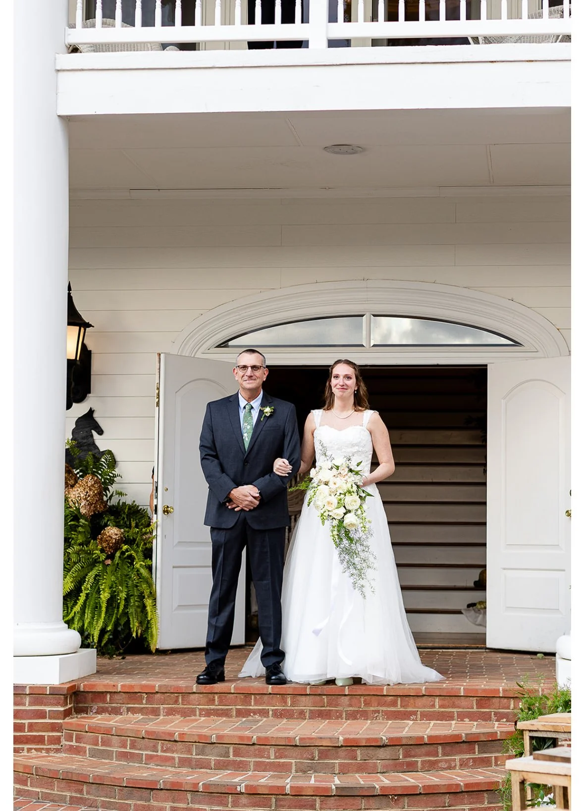 A bride in a white wedding dress holding a bouquet, standing next to a man in a dark suit on the porch of a house, likely during a wedding ceremony.