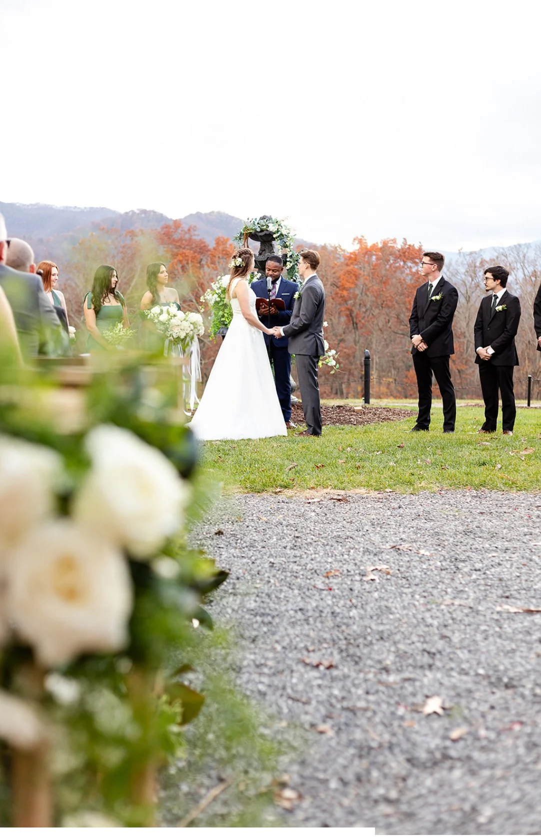 A wedding ceremony outdoors with the bride and groom holding hands, officiant reading vows, bridesmaids on the left, groomsmen on the right, with autumn trees and hills in the background.