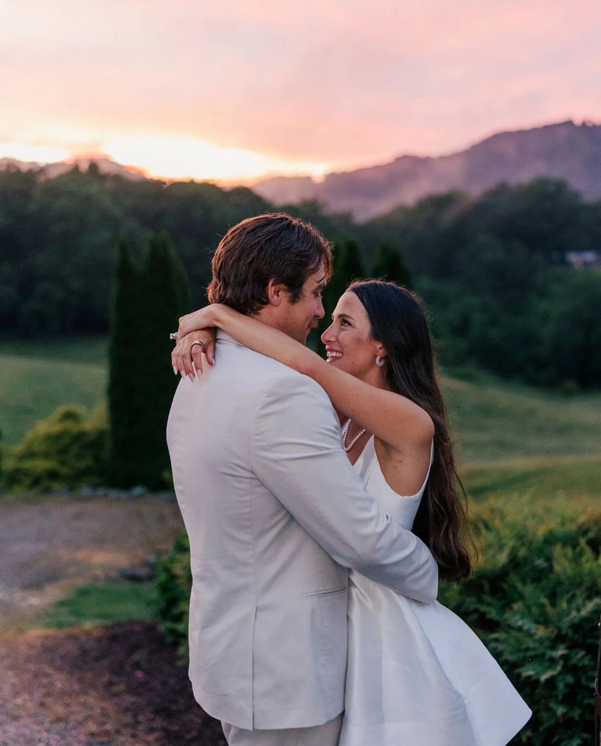 A couple dressed in white embracing outdoors during sunset, with mountains and trees in the background.