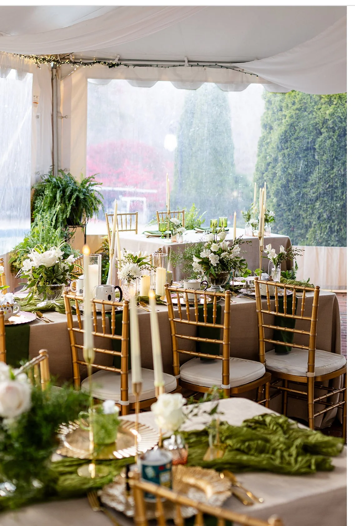 A decorated outdoor wedding or event reception area with gold chiavari chairs, floral centerpieces in vases, tall candles, and greenery, set inside a white tent with clear plastic walls and string lights.