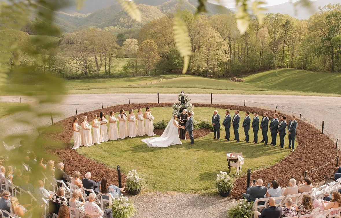 Outdoor wedding ceremony with bride and groom exchanging vows, surrounded by bridesmaids and groomsmen, on a grassy area with a large floral arrangement and guests seated nearby in scenic nature setting.