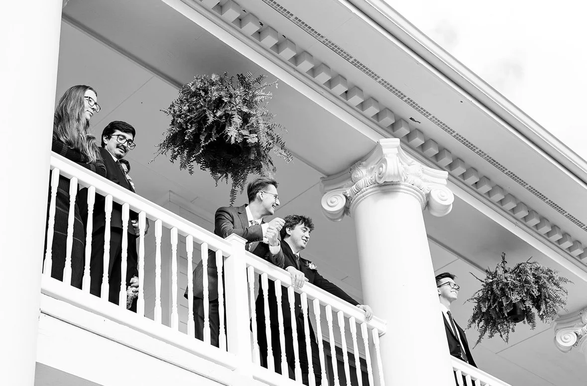 A group of five people on a balcony with white railing, decorated with hanging plants, in a building with classical columns and detailed architectural moldings.