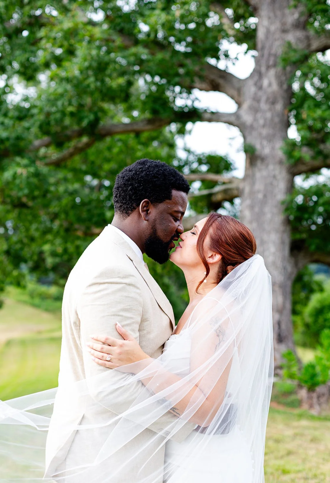 A newlywed couple, a Black man and a White woman with red hair, is embracing outdoors in front of a large tree. The woman is wearing a white wedding dress and veil, and the man is dressed in a beige suit. They are about to kiss, with their faces clos