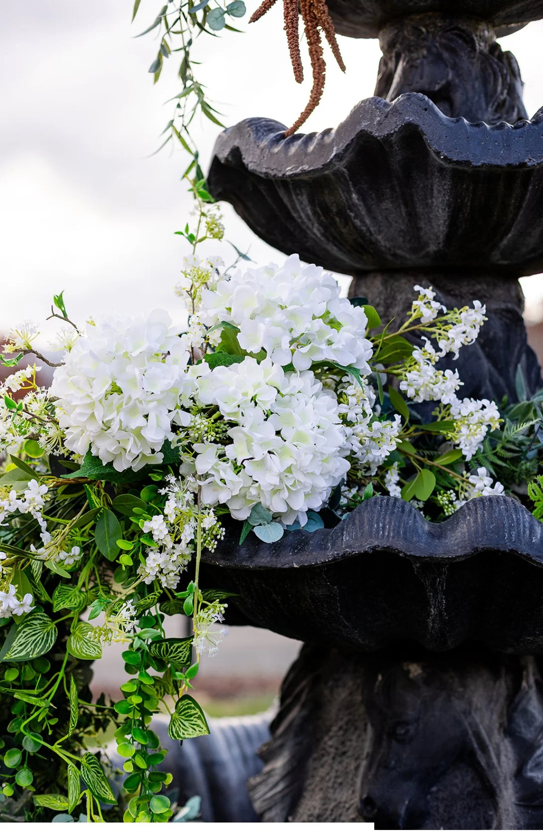 Close-up of a decorative black fountain with white hydrangea flowers and greenery cascading from its upper basin, against a cloudy sky background.