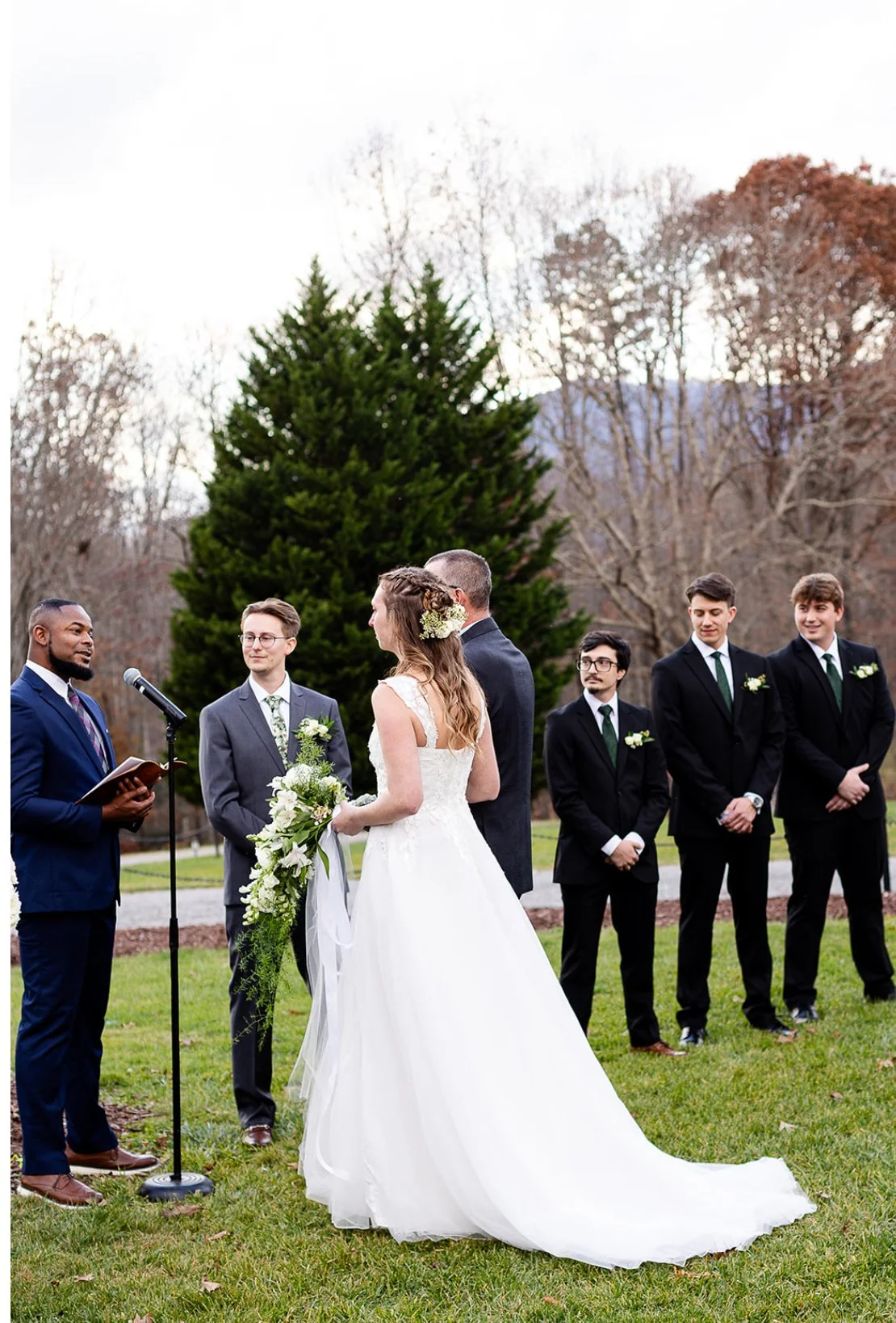 A wedding ceremony outdoors with the bride and groom facing each other and exchange vows. The bride wears a white gown and holds a bouquet, while the groom is in a dark suit. Several groomsmen in dark suits stand to the right, and a man on the left o