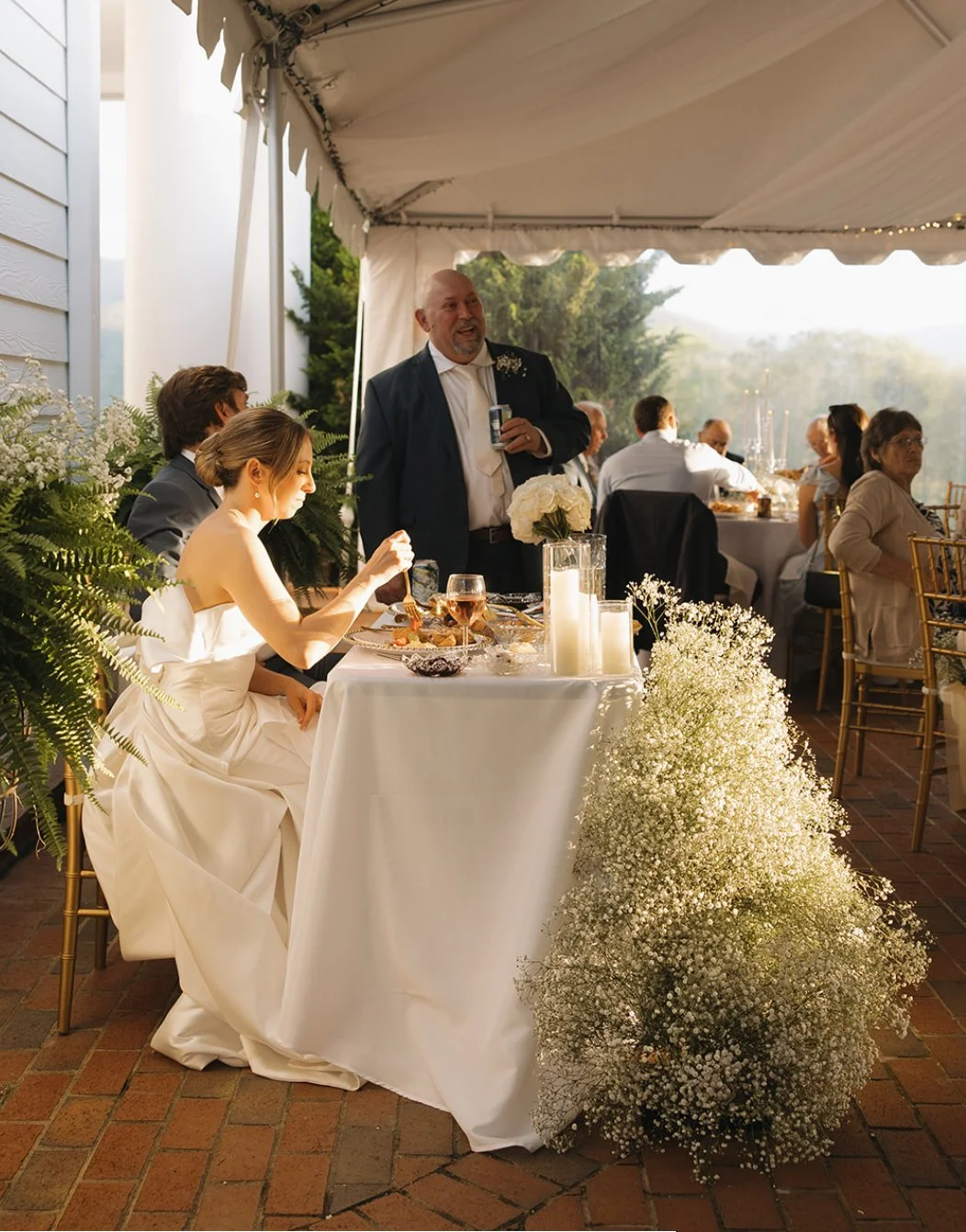 A wedding reception under a tent with a bride in a white gown seated at a table, a groom in a gray suit, and a man giving a toast holding a drink. The table has white flowers, candles, and food. The background includes other guests sitting at tables 