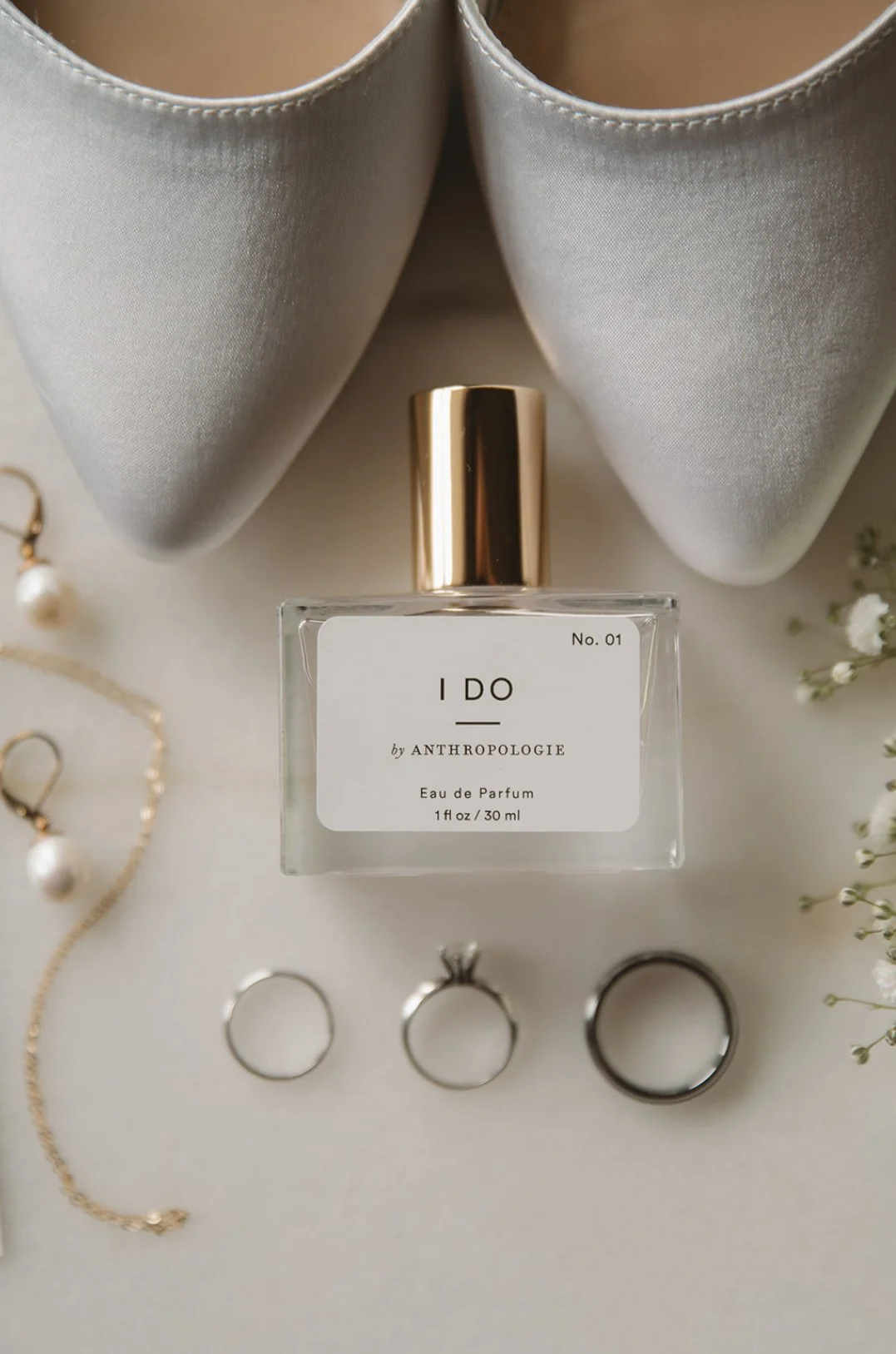 A perfume bottle labeled 'I DO by ANTHROPOLOGIE, Eau de Parfum, 30 ml' surrounded by jewelry, white shoes, and flowers.