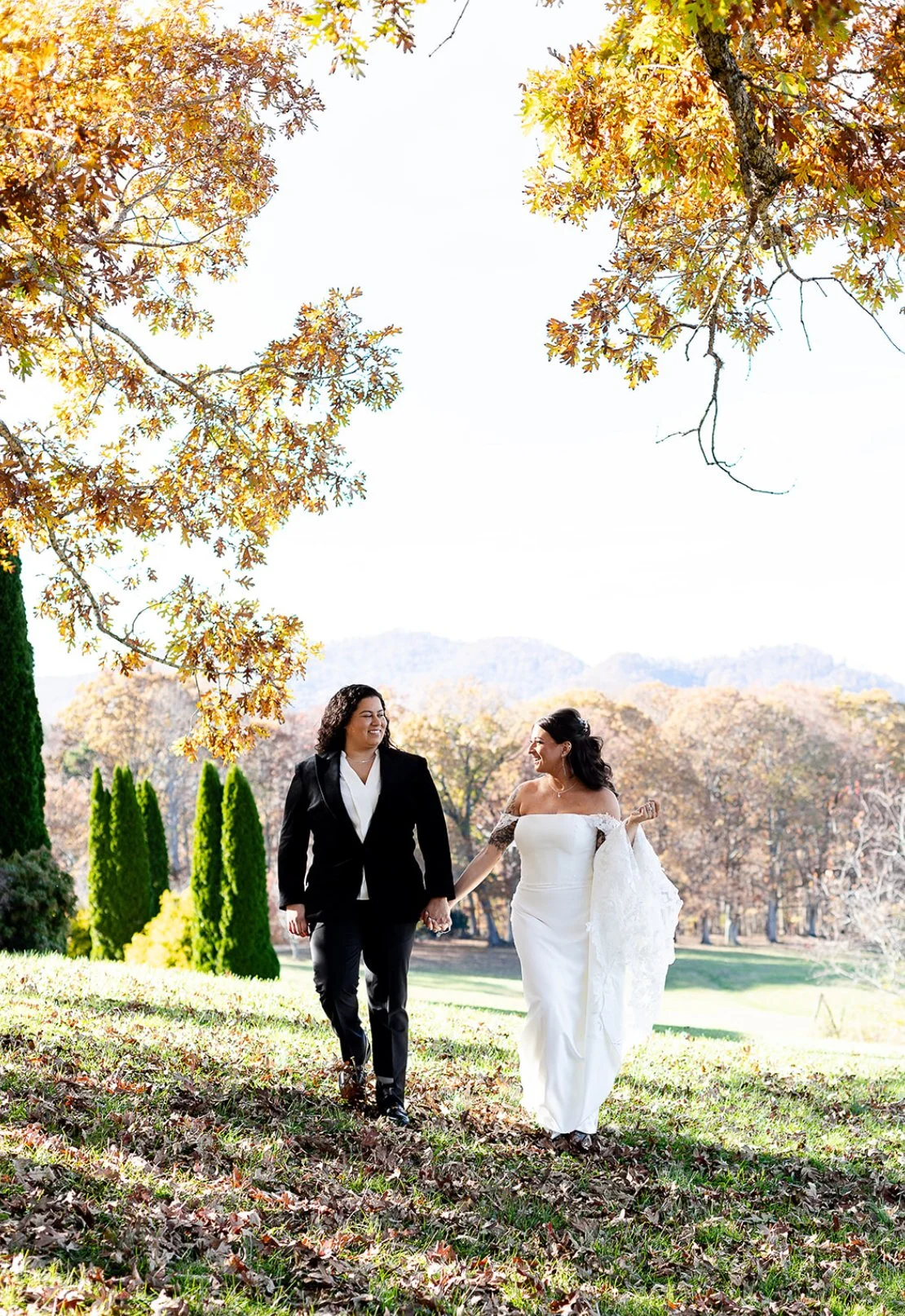 Two women, one in a wedding dress and the other in a black suit, holding hands and walking outdoors in a park with autumn trees and mountains in the background.