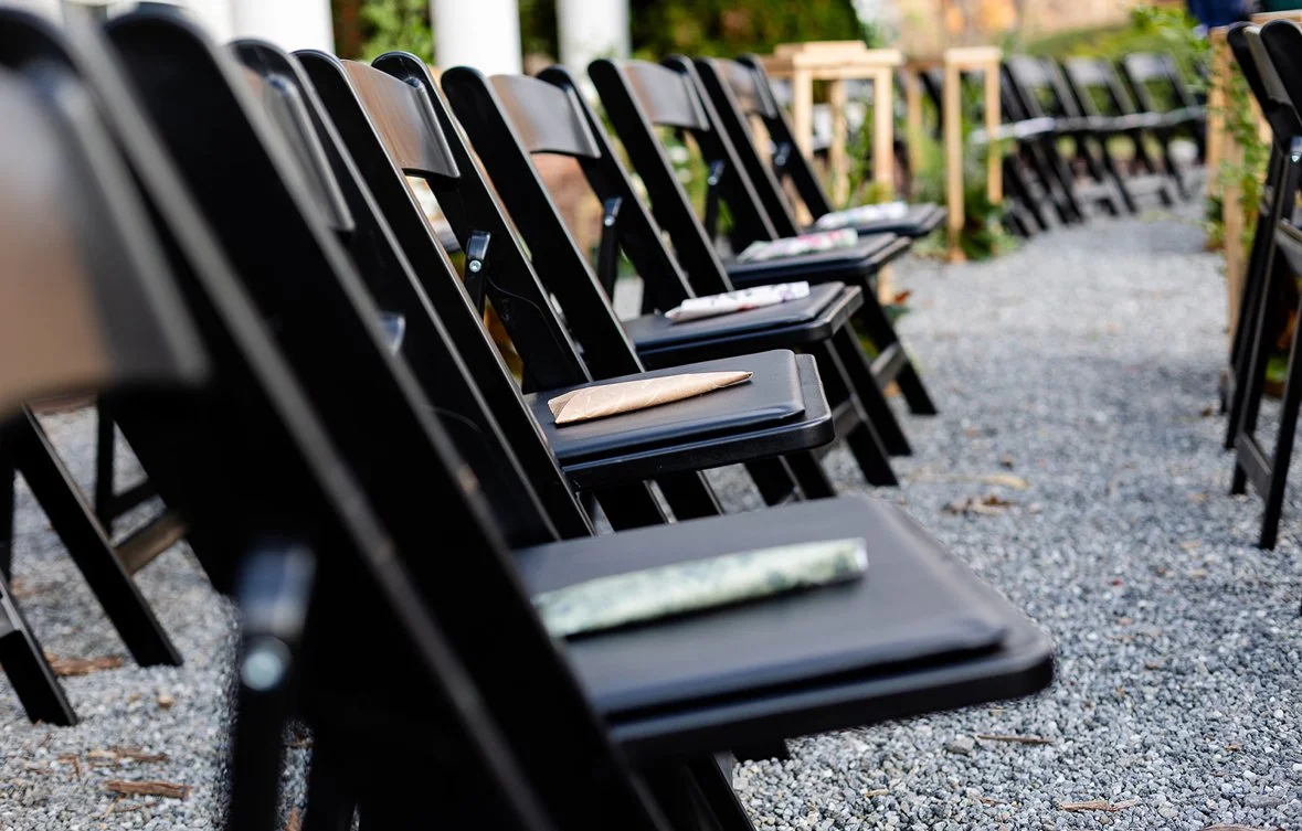 Black folding chairs arranged outdoors on a gravel surface, each with a wrapped napkin or paper on the seat.