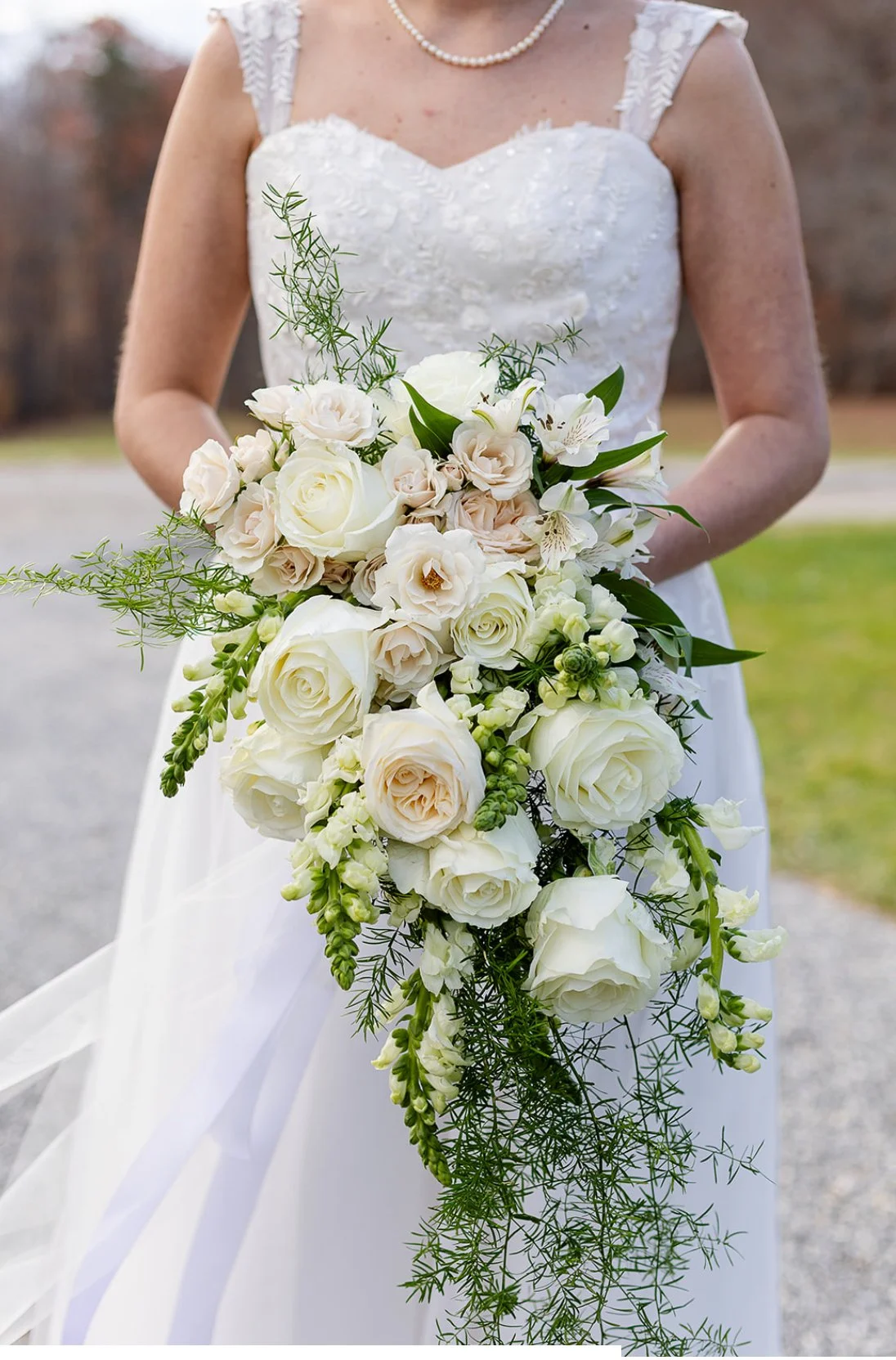 Bride holding a cascading bouquet of white roses, lilies, and greenery in an outdoor setting.