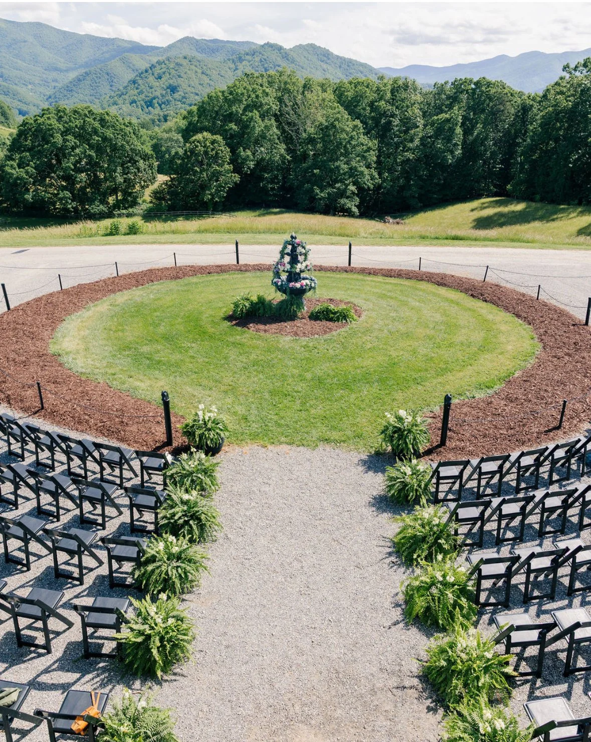 Outdoor wedding or event setup with black chairs arranged in rows on gravel, facing a green grassy area with a decorated water fountain in the center, surrounded by a circular flower bed and a backdrop of trees and mountains.