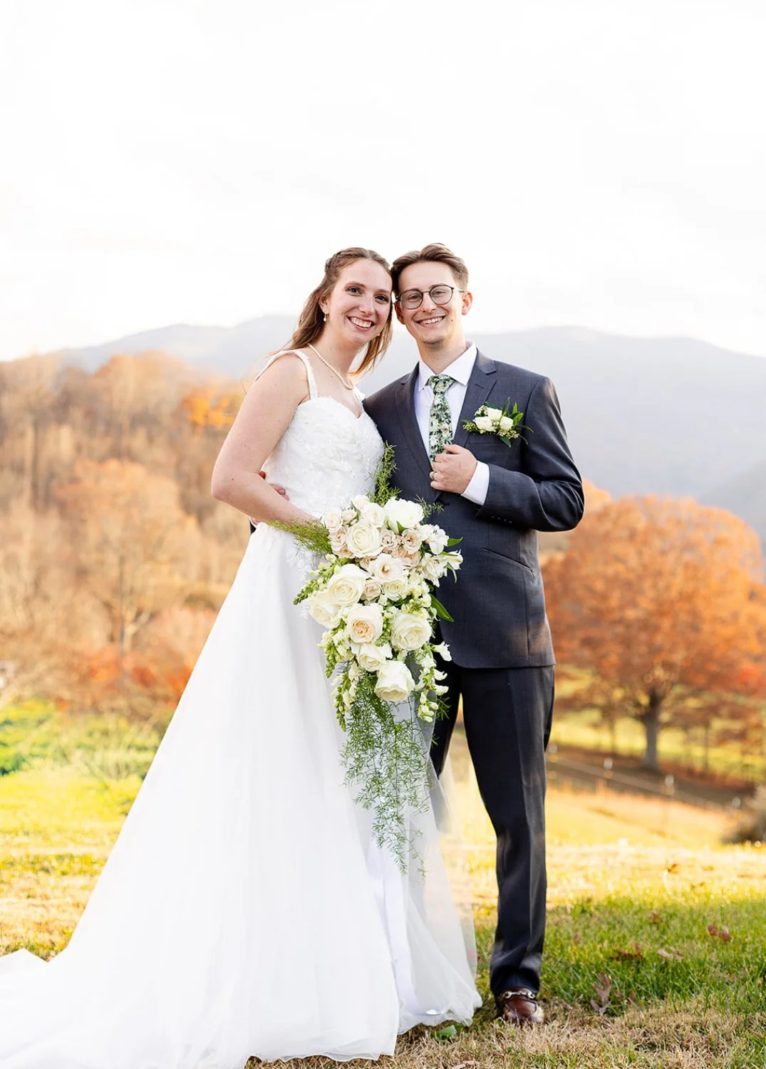 A bride and groom standing outdoors with mountains and trees with fall foliage in the background, smiling and posing for their wedding photo. The bride is holding a large bouquet of white roses and greenery, and the groom is wearing a dark suit with 