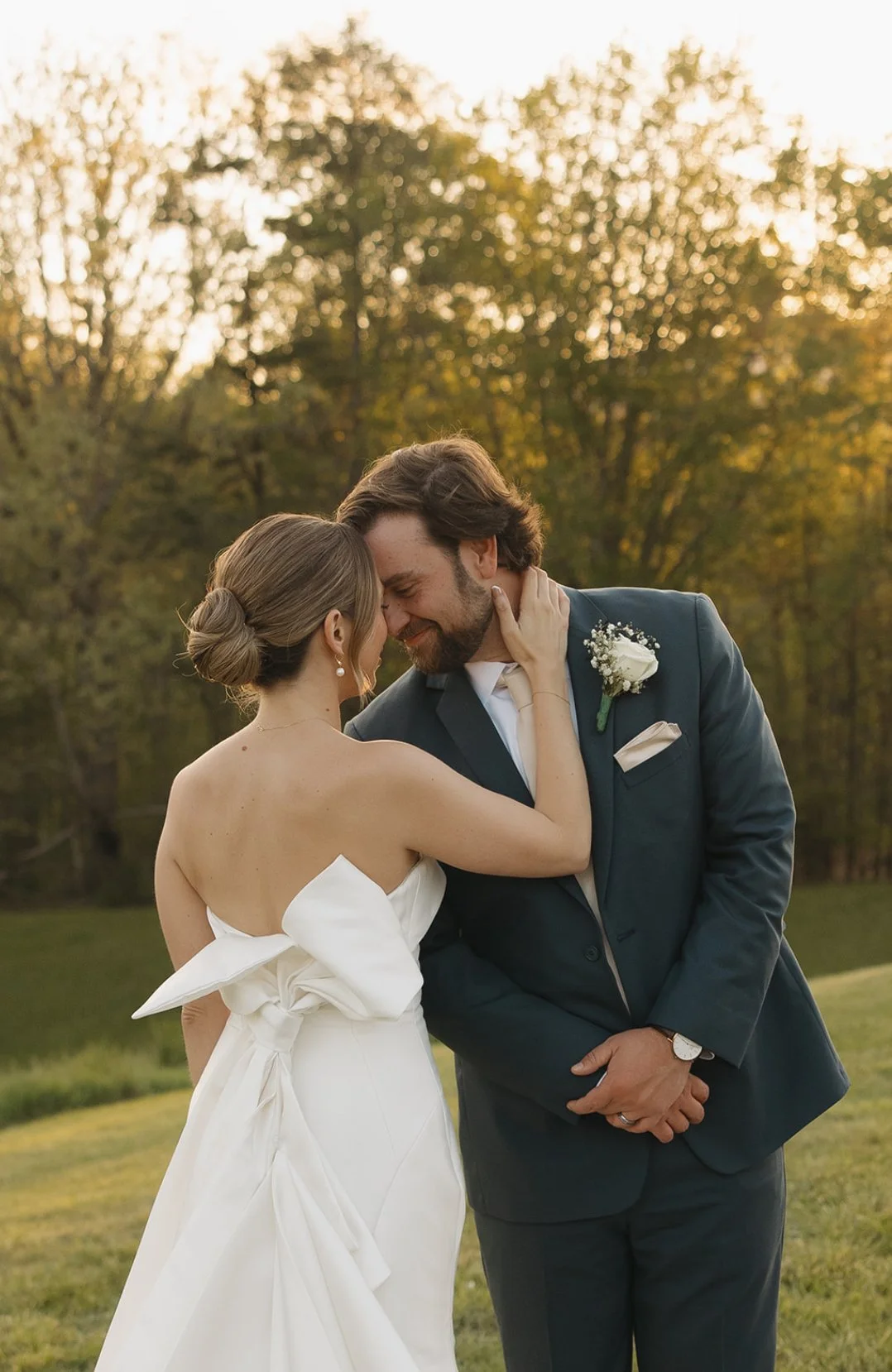 A bride and groom share a close, intimate moment outdoors at sunset, with trees in the background. The bride is wearing a white wedding dress with a large bow on the back, and the groom is in a dark suit with a boutonniere. They are touching forehead