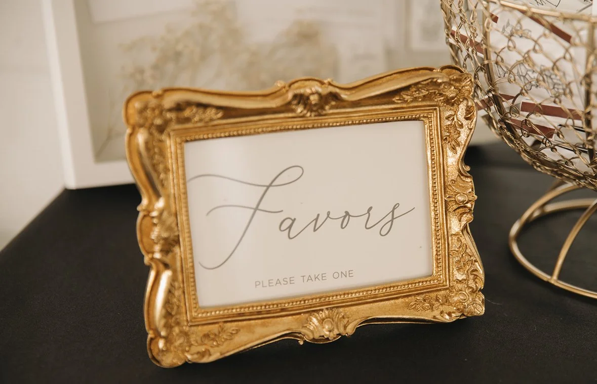 Gold ornate picture frame with a white sign inside that reads 'Favorites' in cursive font and 'Please take one' in smaller print, placed on a black table.