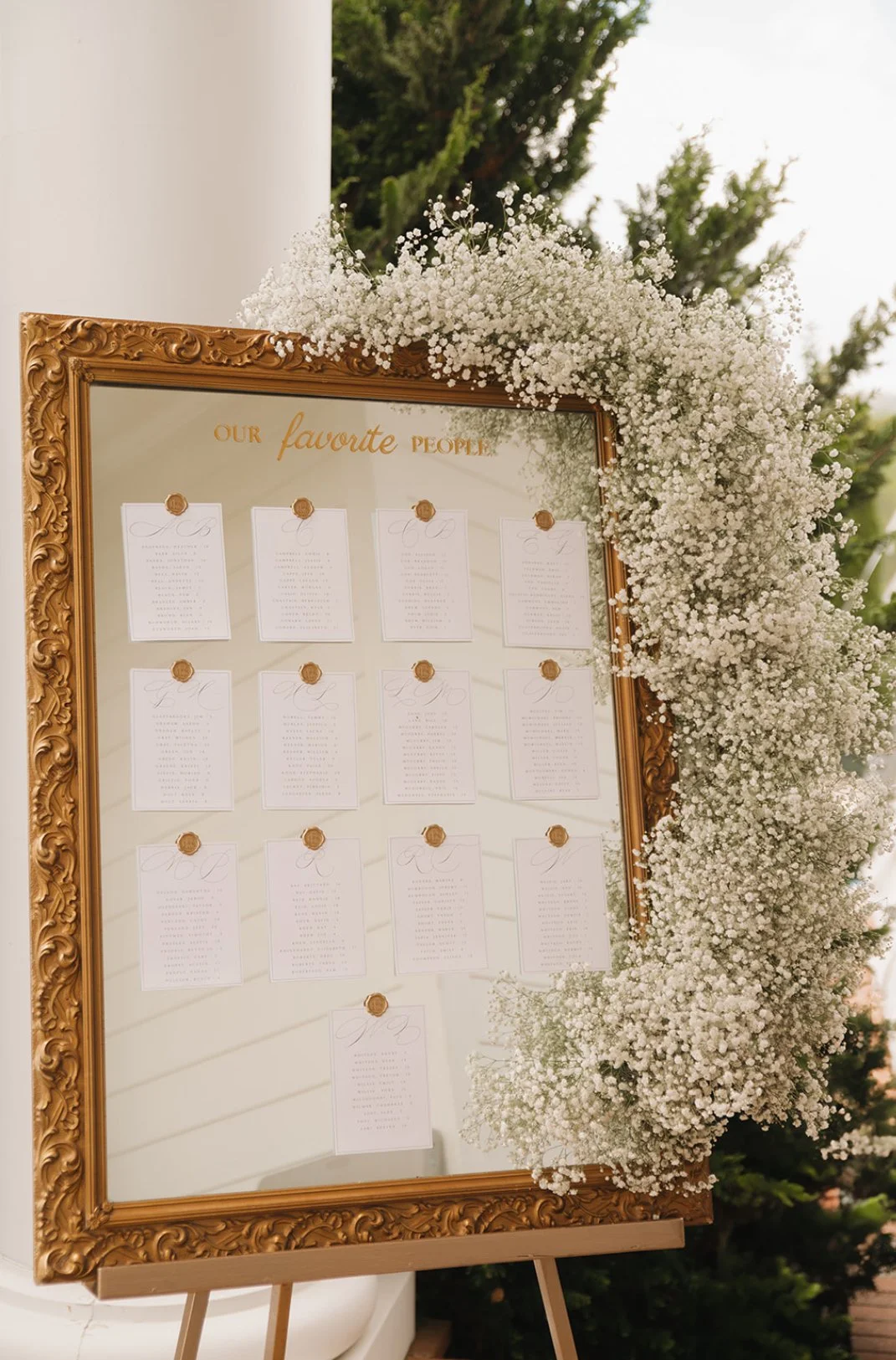 A decorative seating chart display for a wedding, framed in ornate gold and bordered with white baby's breath flowers.