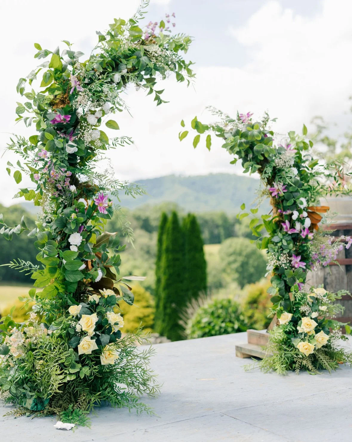 Flower arch decorated with green leaves, white, pink, and purple flowers, set outdoors with trees and hills in the background.