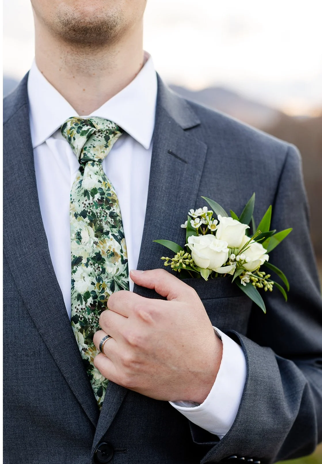 Close-up of a man wearing a gray suit, floral tie, white shirt, with a boutonnière of white roses and greenery on his jacket lapel, holding his lapel with his left hand.