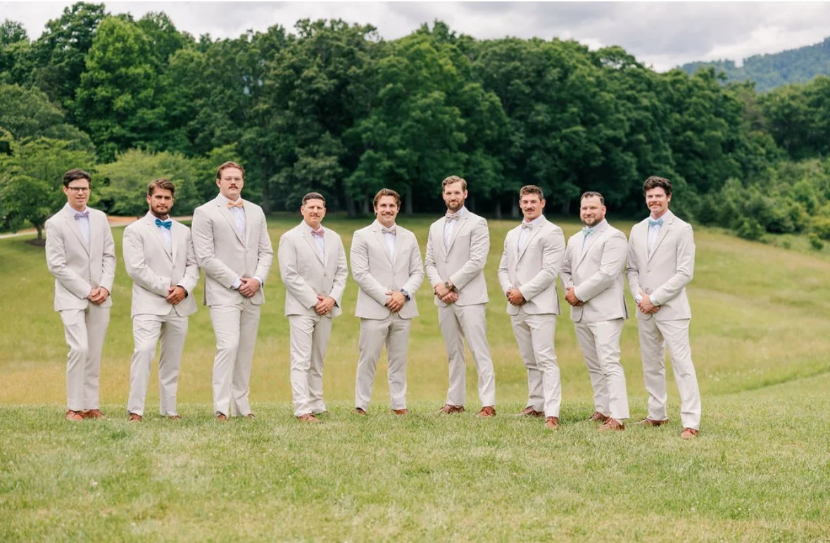 Group of men in white suits standing outdoors on a grassy field with trees in the background.