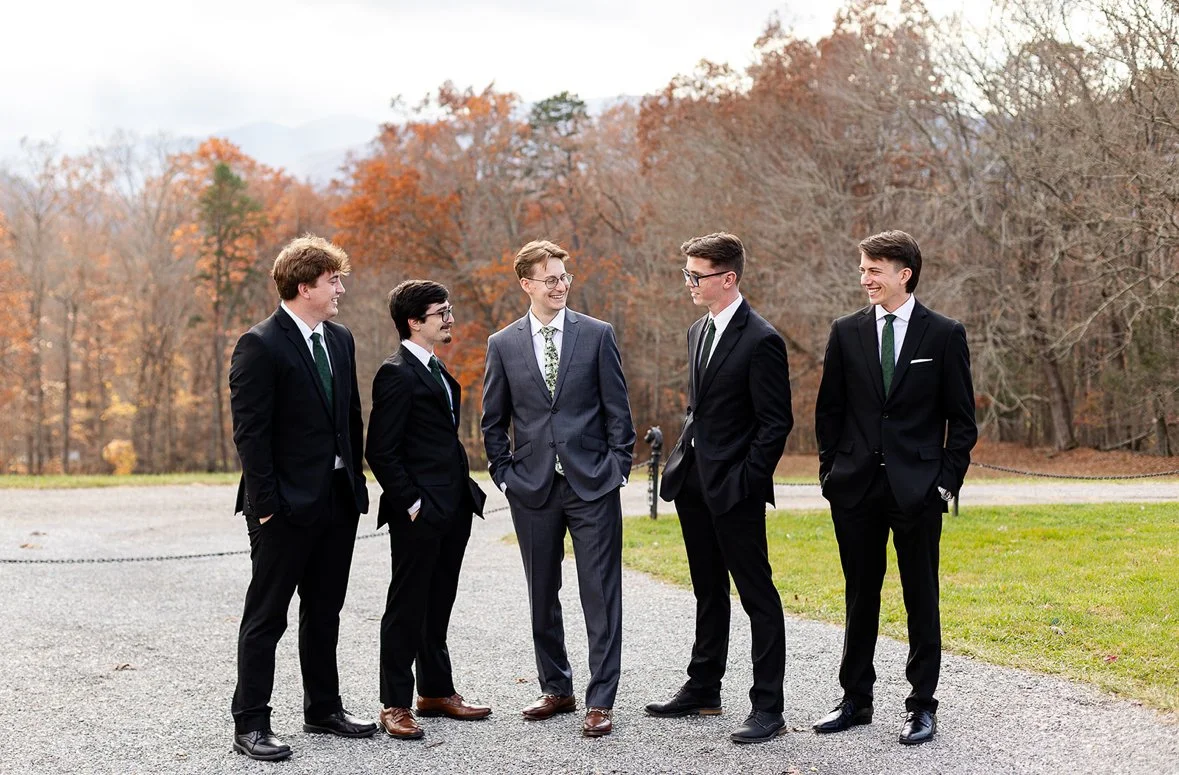 Five young men dressed in suits standing outdoors in a line, engaging in conversation, with autumn trees in the background.