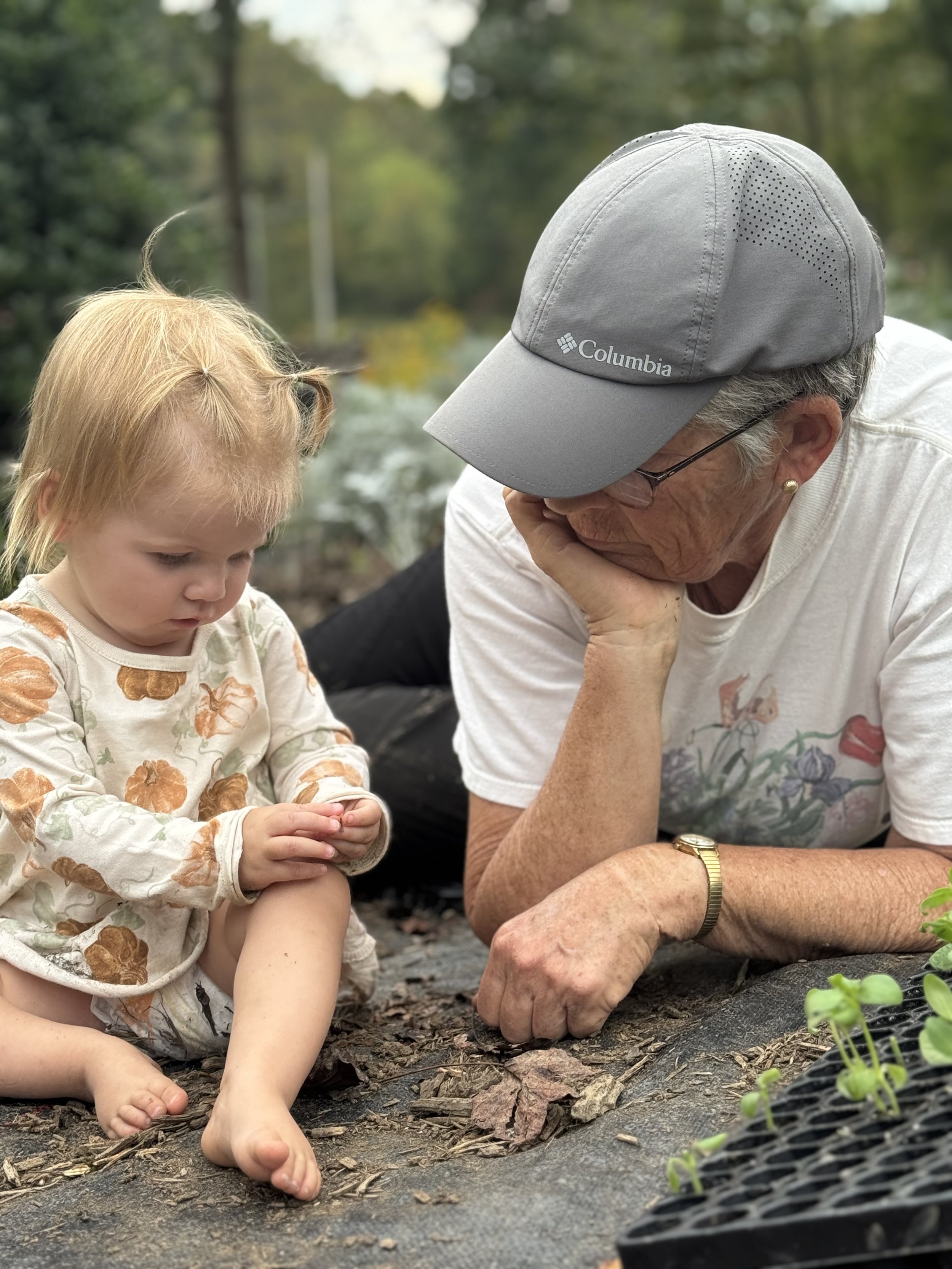 An elderly woman and young girl examining soil and small plants outdoors on a garden bed, with trees and greenery in the background.