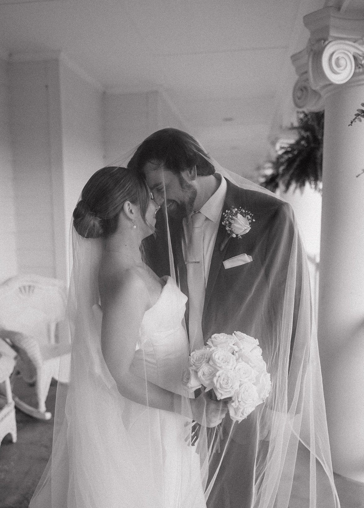 Black and white photo of a bride and groom sharing a moment, with the bride holding a bouquet of roses and both leaning their foreheads together.