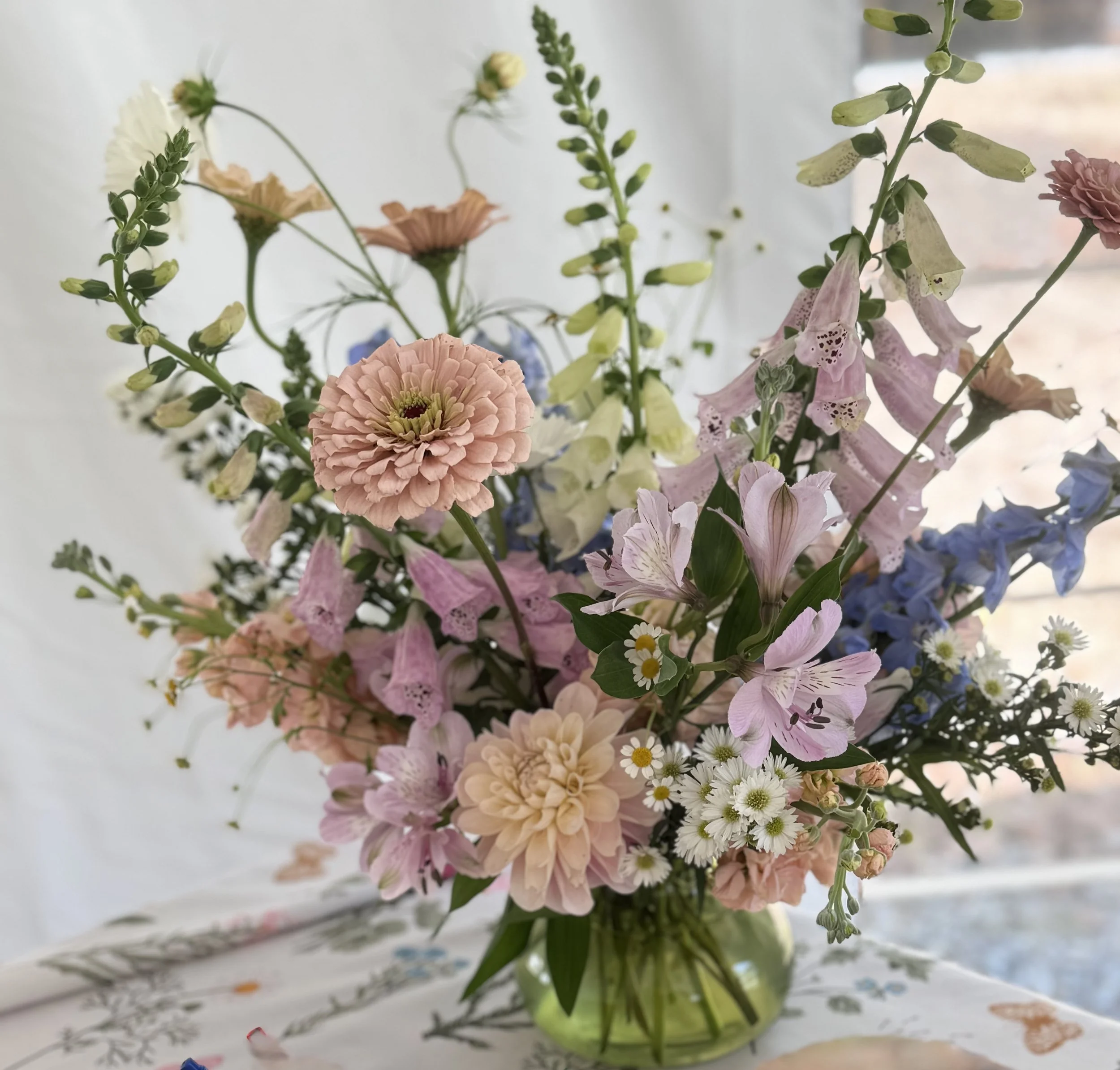 A vibrant bouquet of pastel-colored flowers in a green glass vase on a table with a white embroidered cloth.