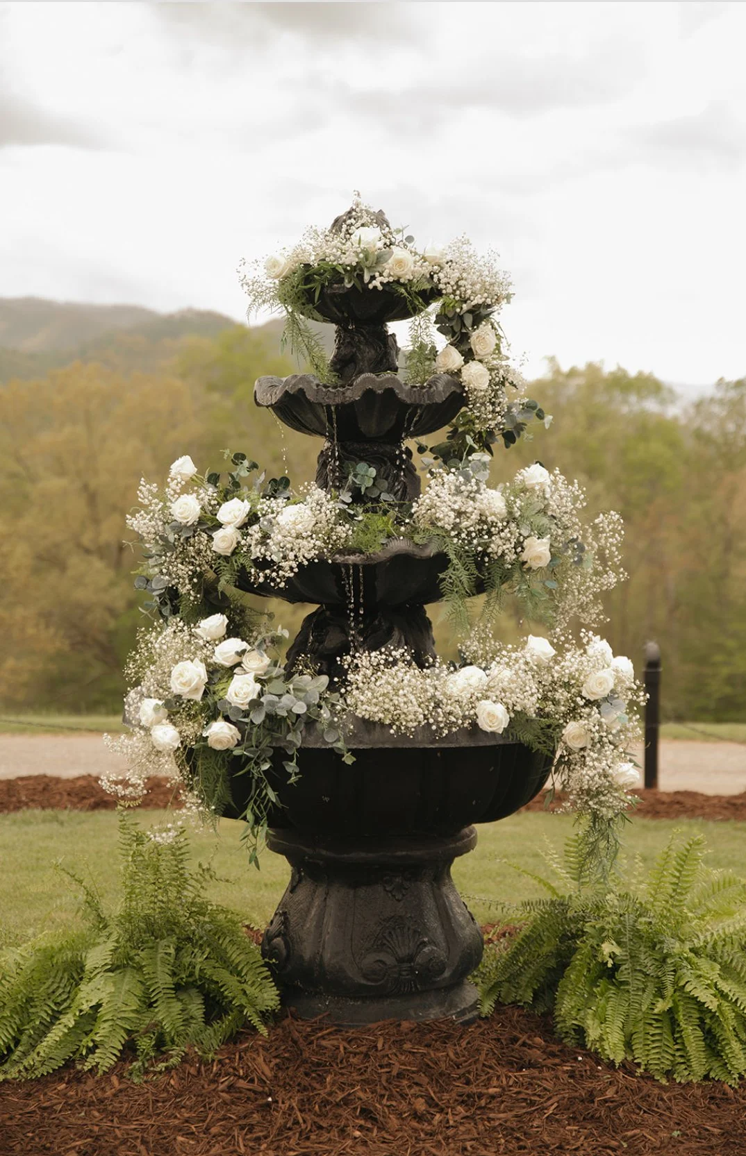 A three-tier black fountain decorated with white roses, baby's breath, and greenery, set outdoors against a cloudy sky and distant trees.