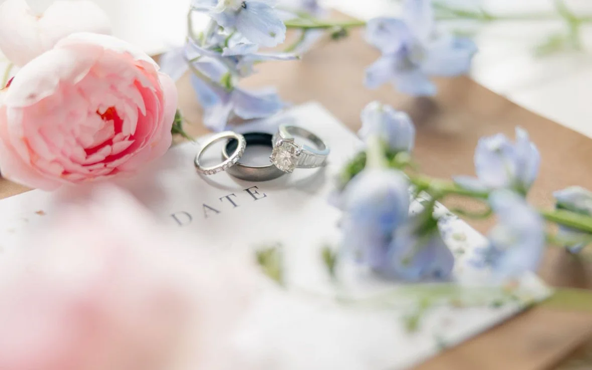 Wedding rings placed on a handwritten date card surrounded by pink and blue flowers.