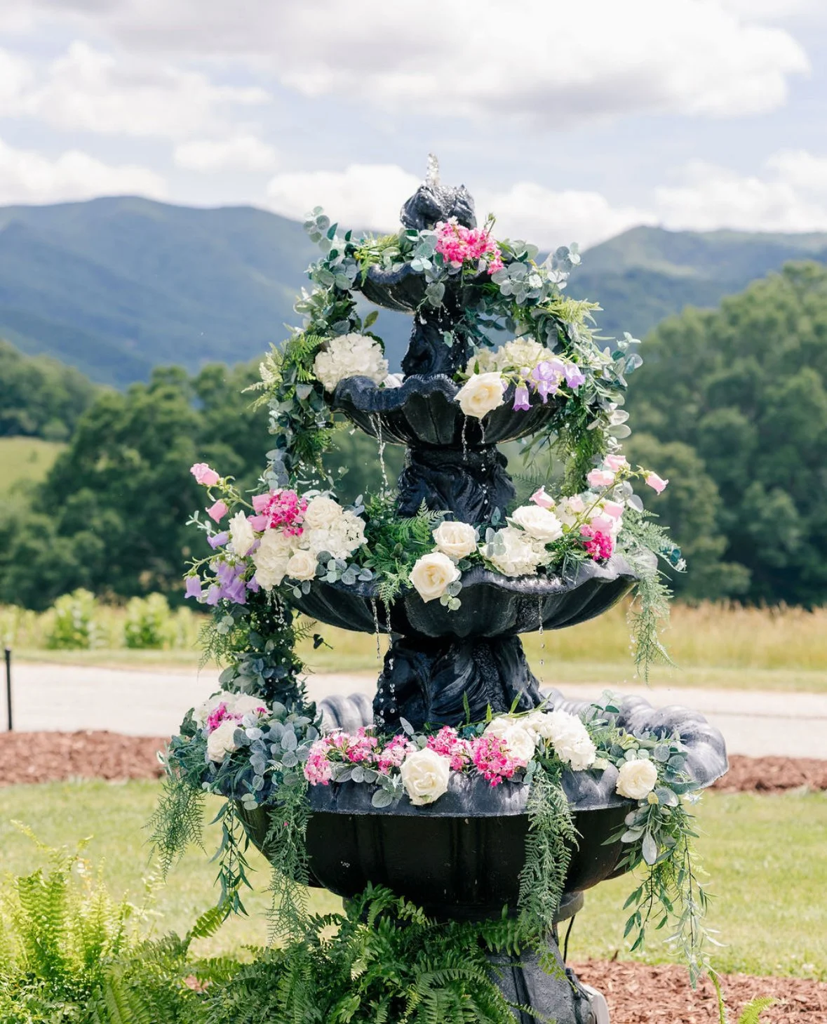 A black multi-tiered outdoor fountain decorated with pink, white, and purple flowers and green foliage, set against a mountain landscape with trees and clouds.