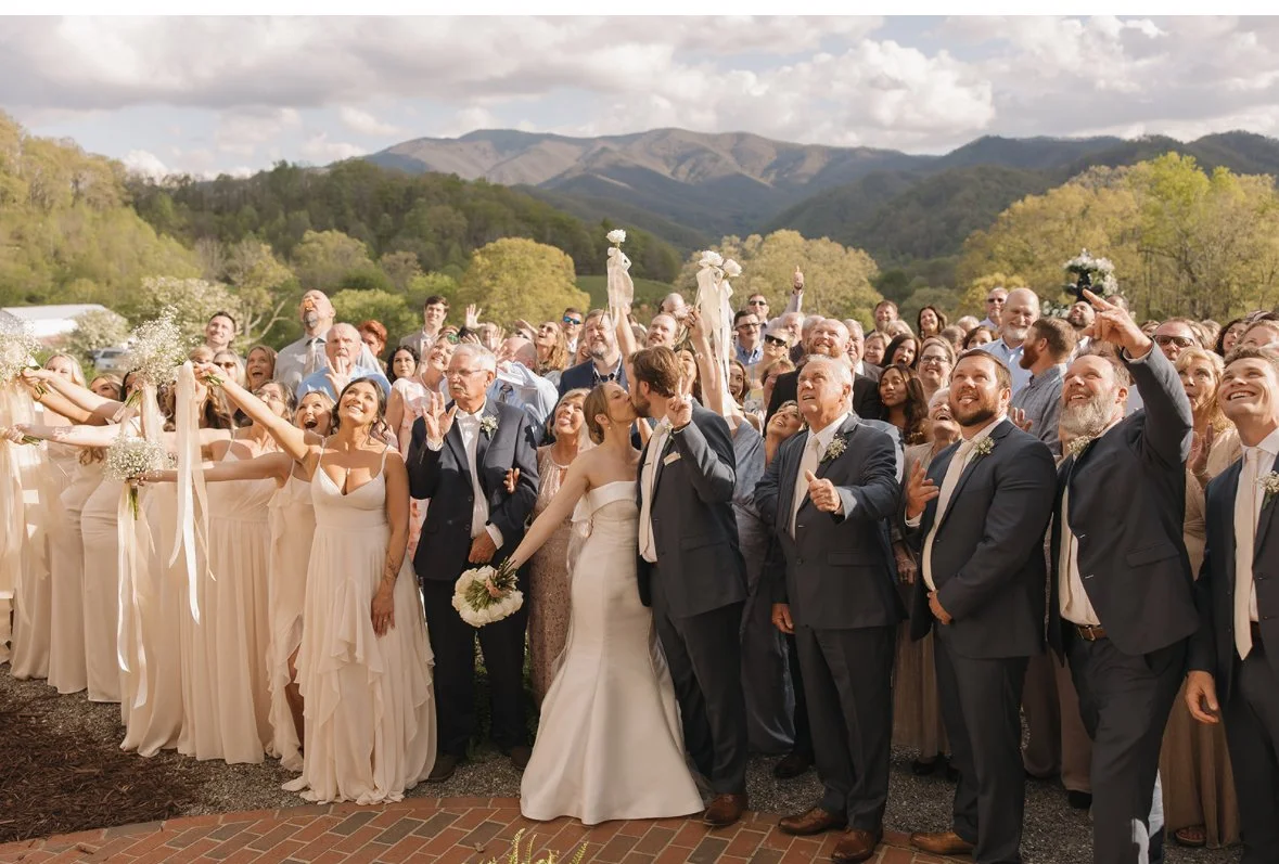 A wedding celebration with a large group of guests outdoors, with mountains and trees in the background, during the daytime.