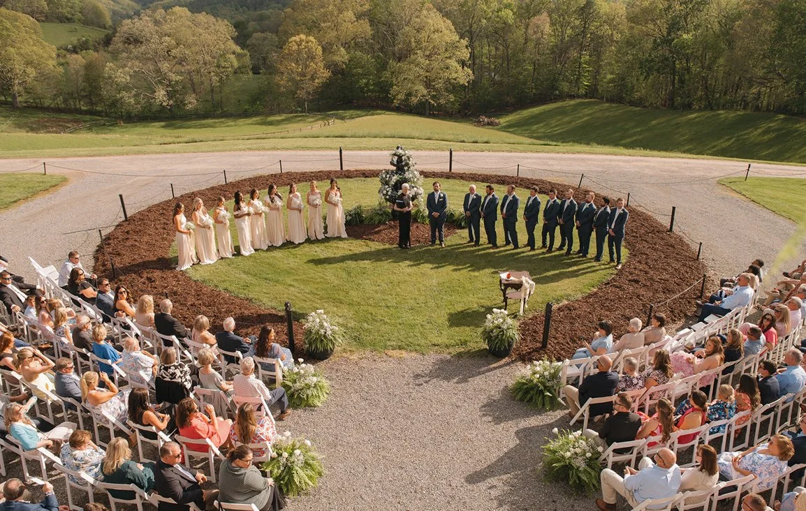 Outdoor wedding ceremony with the bride and groom standing in the center, bridesmaids to the left, groomsmen to the right, guests seated in white chairs facing them, in a lush green landscape with trees and hills in the background.