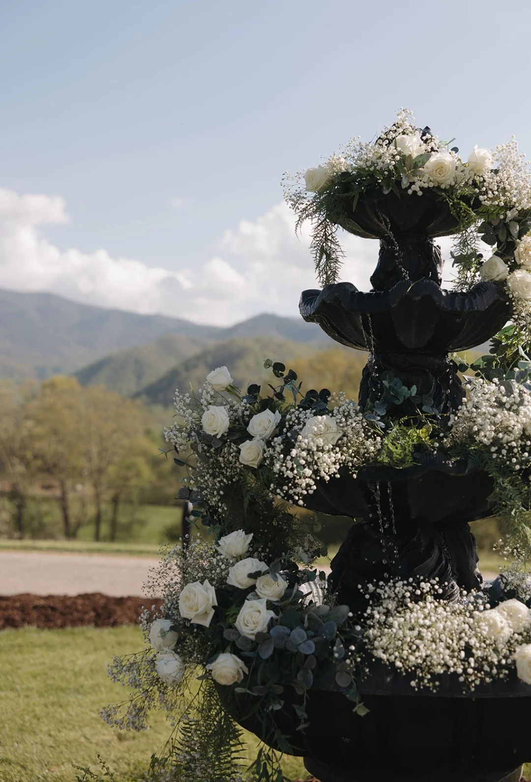 A black multi-tiered fountain decorated with white roses, baby's breath, and greenery outdoors with mountains and trees in the background.