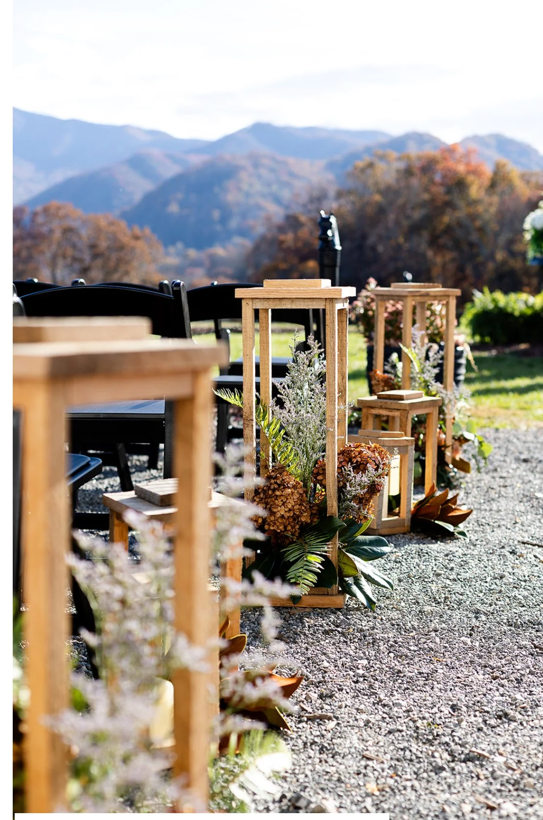 Outdoor wedding or event setup with wooden lanterns and floral arrangements along a gravel path, scenic mountain view with trees in autumn colors in the background.