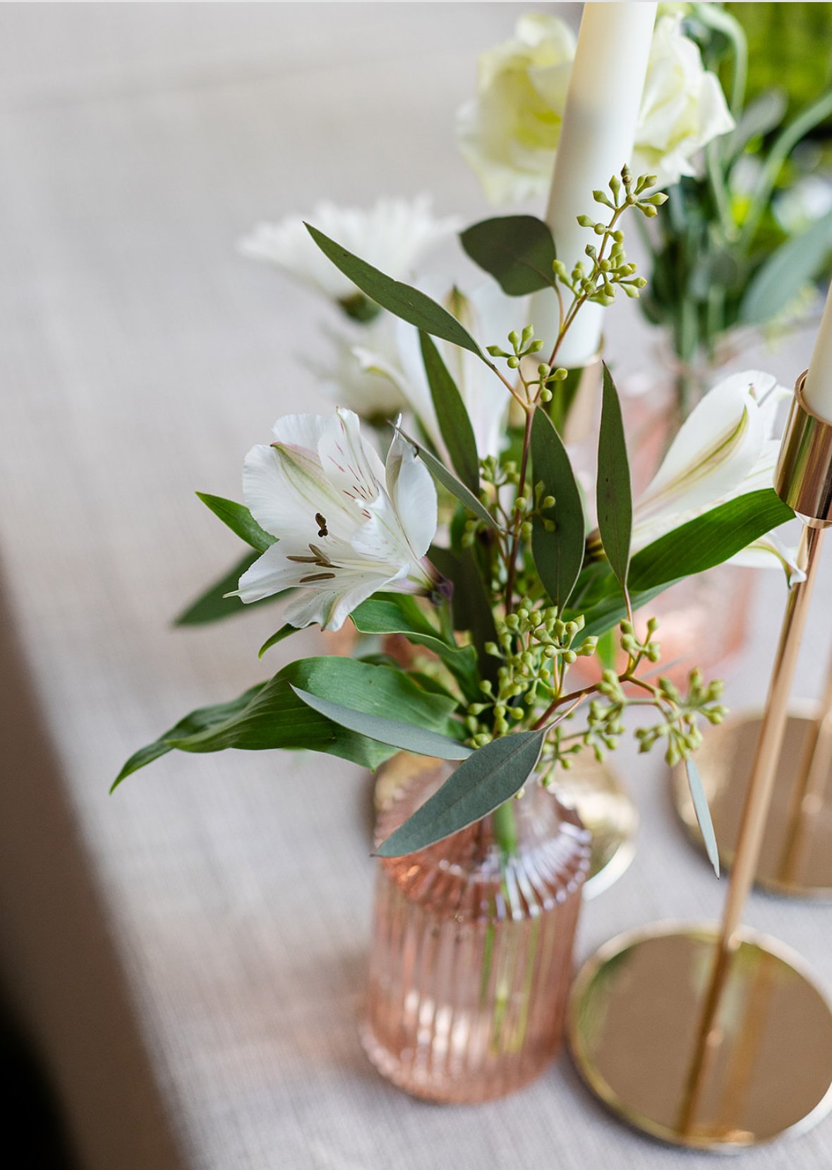 Close-up of a floral arrangement in a pink glass vase, featuring white lilies, eucalyptus leaves, and green buds on a table.