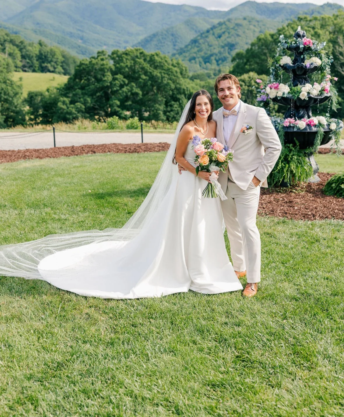 A bride and groom standing on a green lawn with mountains and trees in the background, smiling at the camera, with a decorative floral fountain to their right.