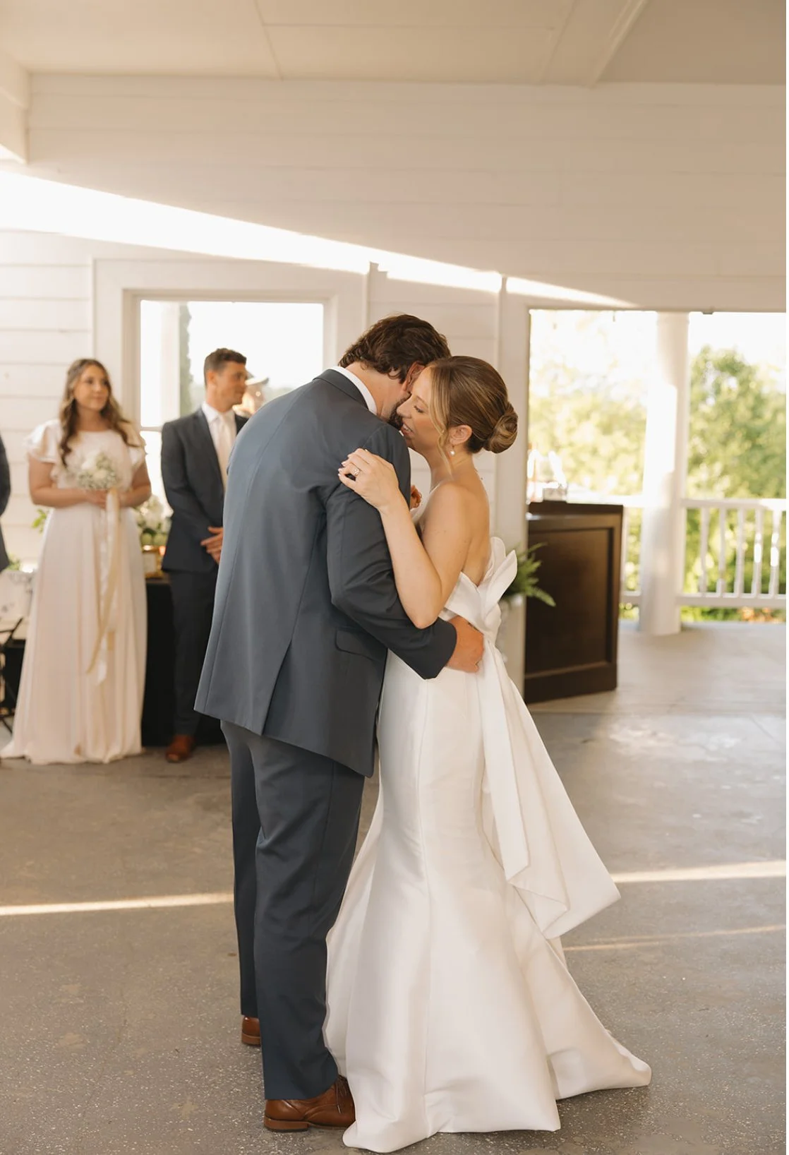 A bride and groom share a dance during their wedding ceremony inside a bright, white-walled venue, with bridesmaids and groomsmen watching in the background.